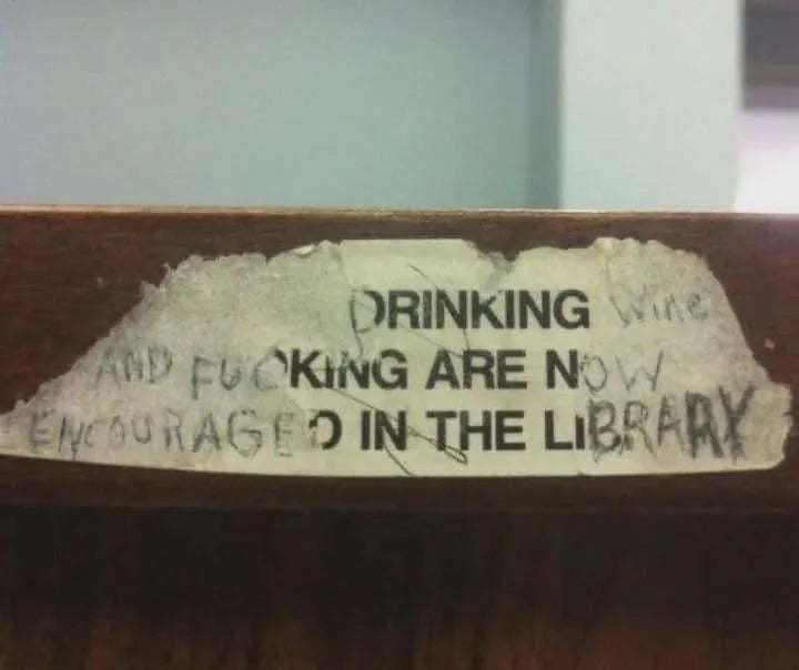 Paper sign with altered text reads: "Drinking wine and fucking are now encouraged in the library."