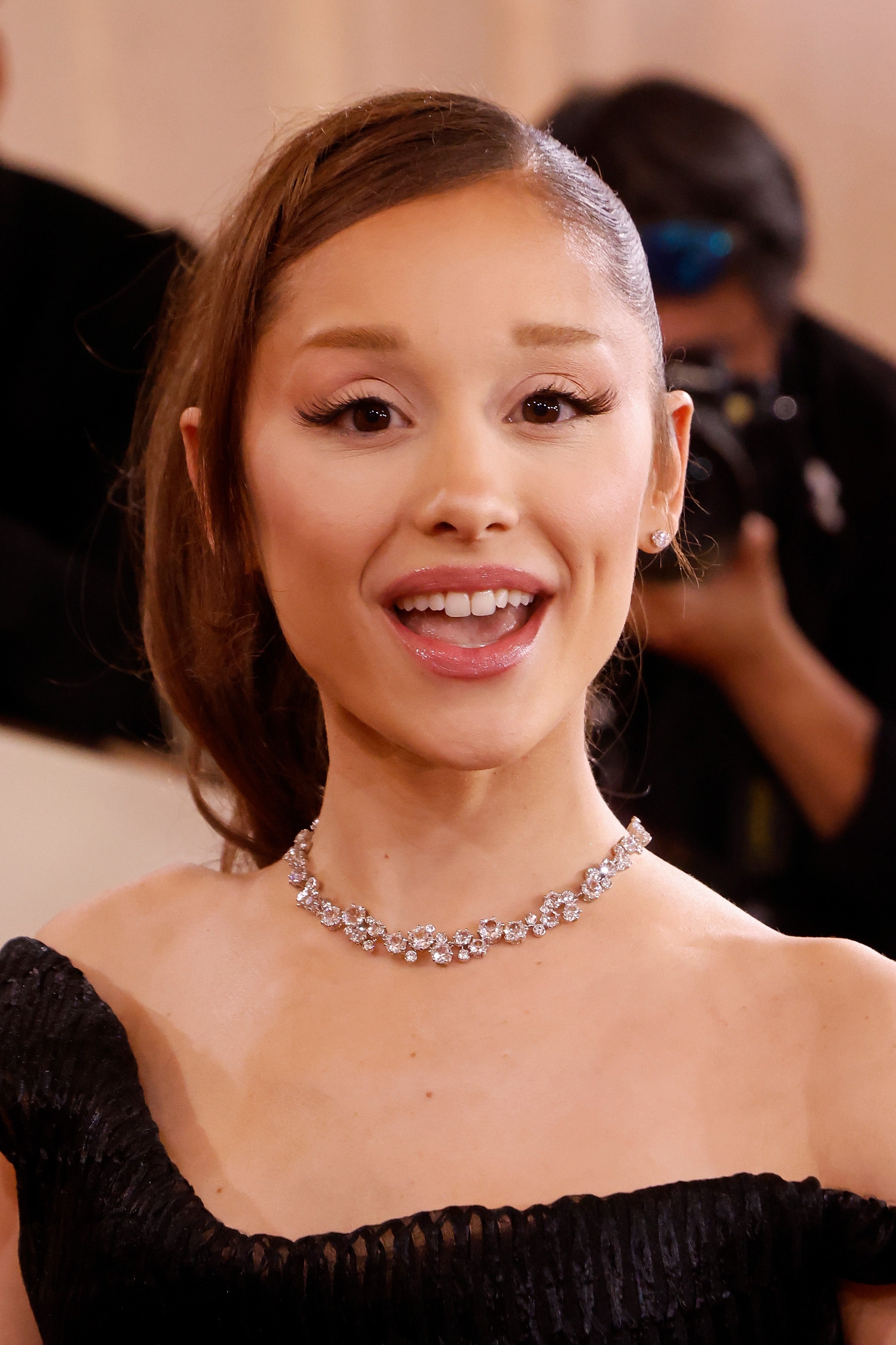 Ariana Grande smiling at an event, wearing a diamond necklace and off-the-shoulder gown