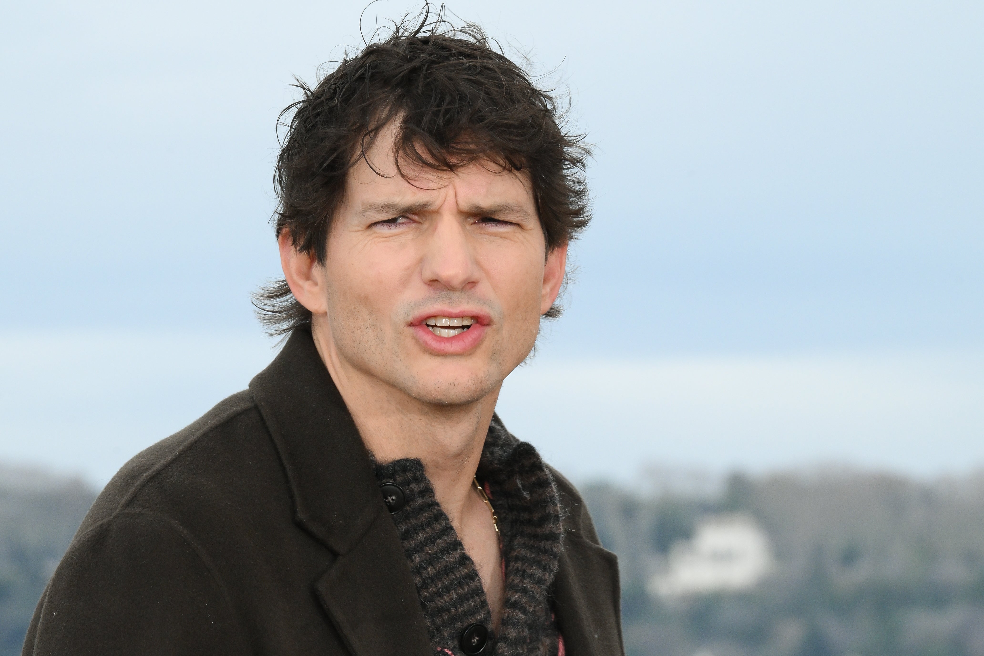 Ashton Kutcher outdoors wearing a dark jacket over a textured shirt, looking at the camera