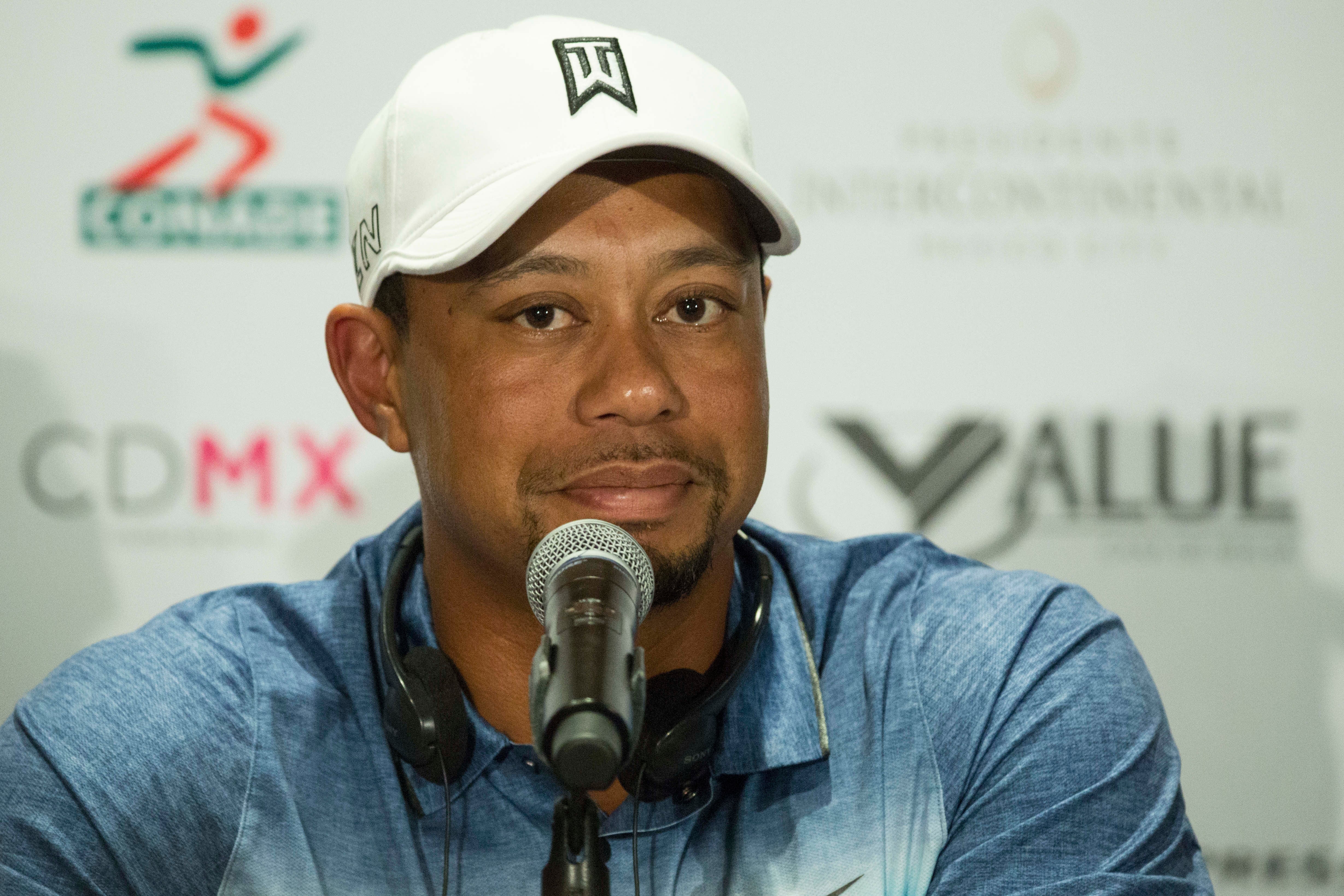 Tiger Woods wearing a cap and casual shirt speaks at a press conference, sitting behind a microphone
