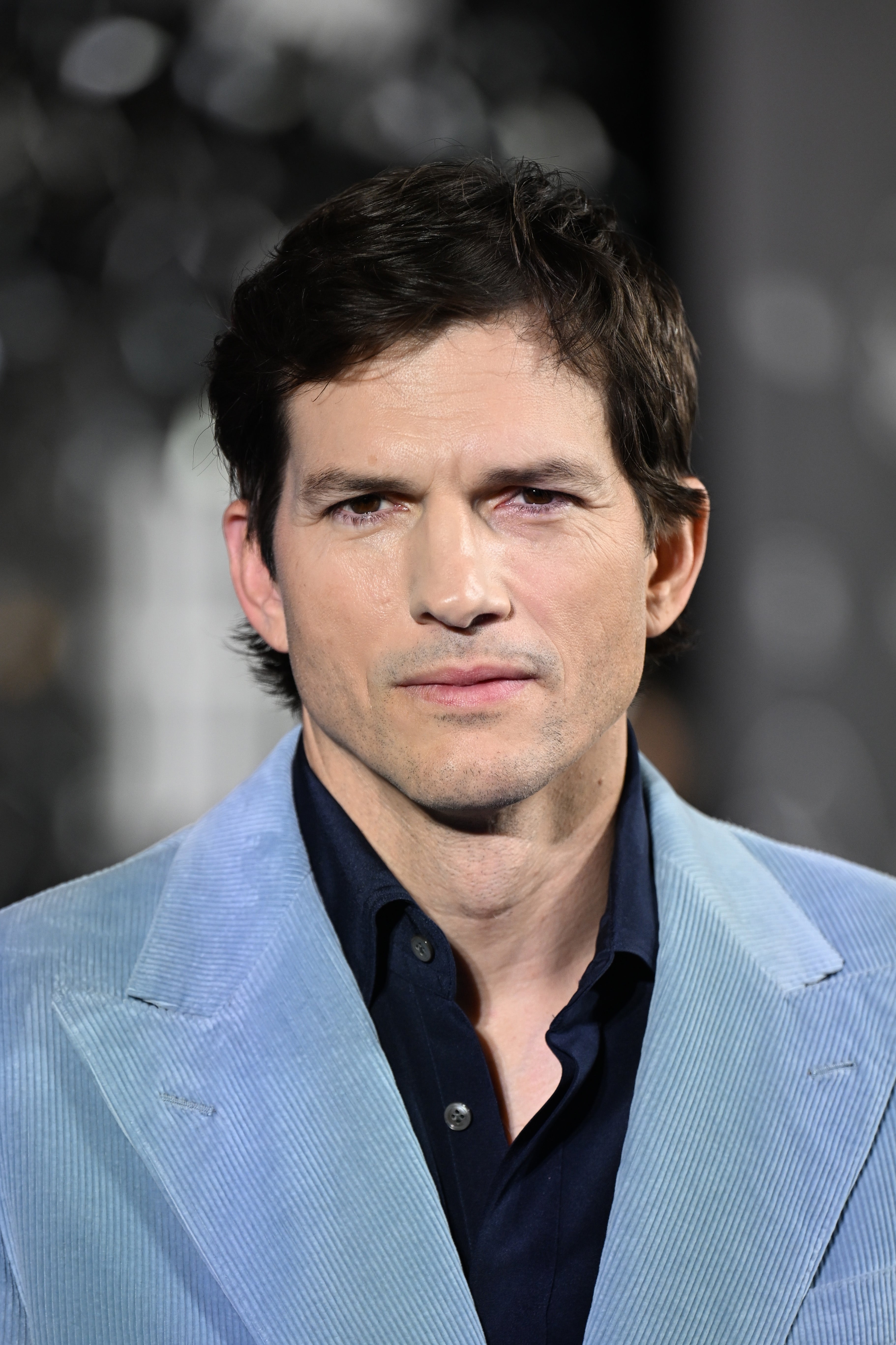 Ashton Kutcher in a formal setting, wearing a textured jacket over a shirt