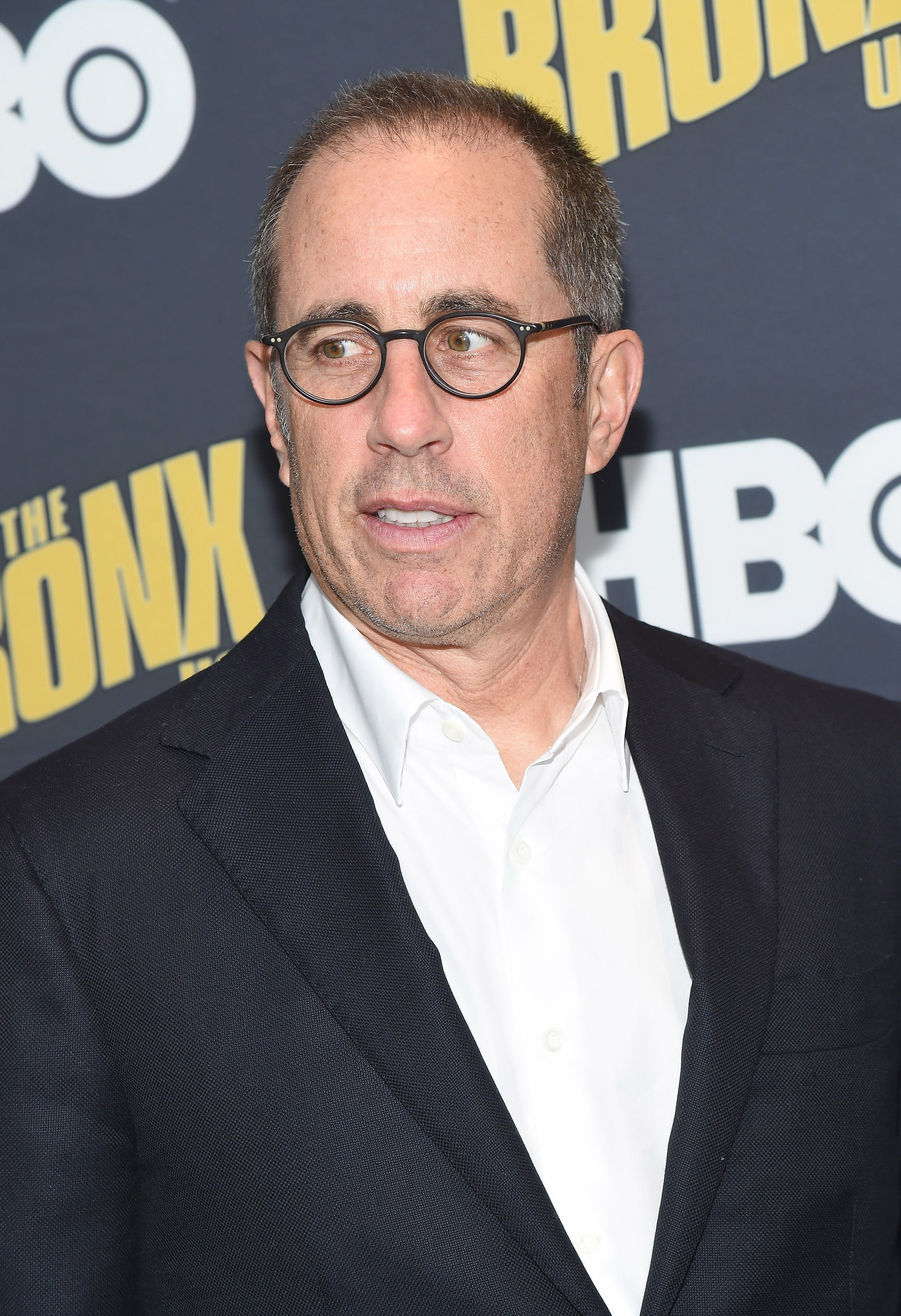 Jerry Seinfeld in glasses, wearing a formal suit, at a media event with a backdrop featuring "Bronx" and "HBO" logos