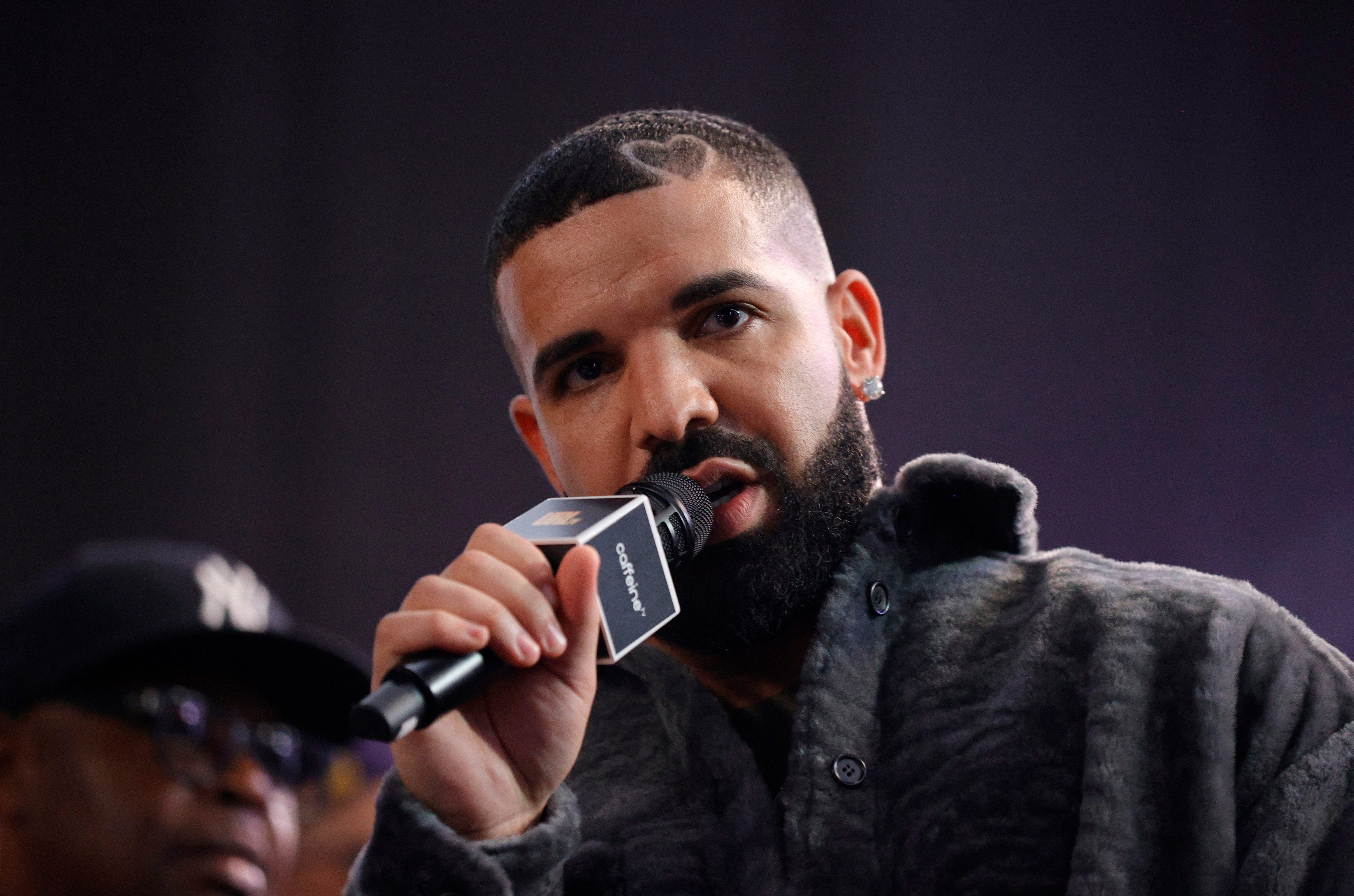 Drake with a well-groomed beard and short hair speaks into a microphone while wearing a textured jacket