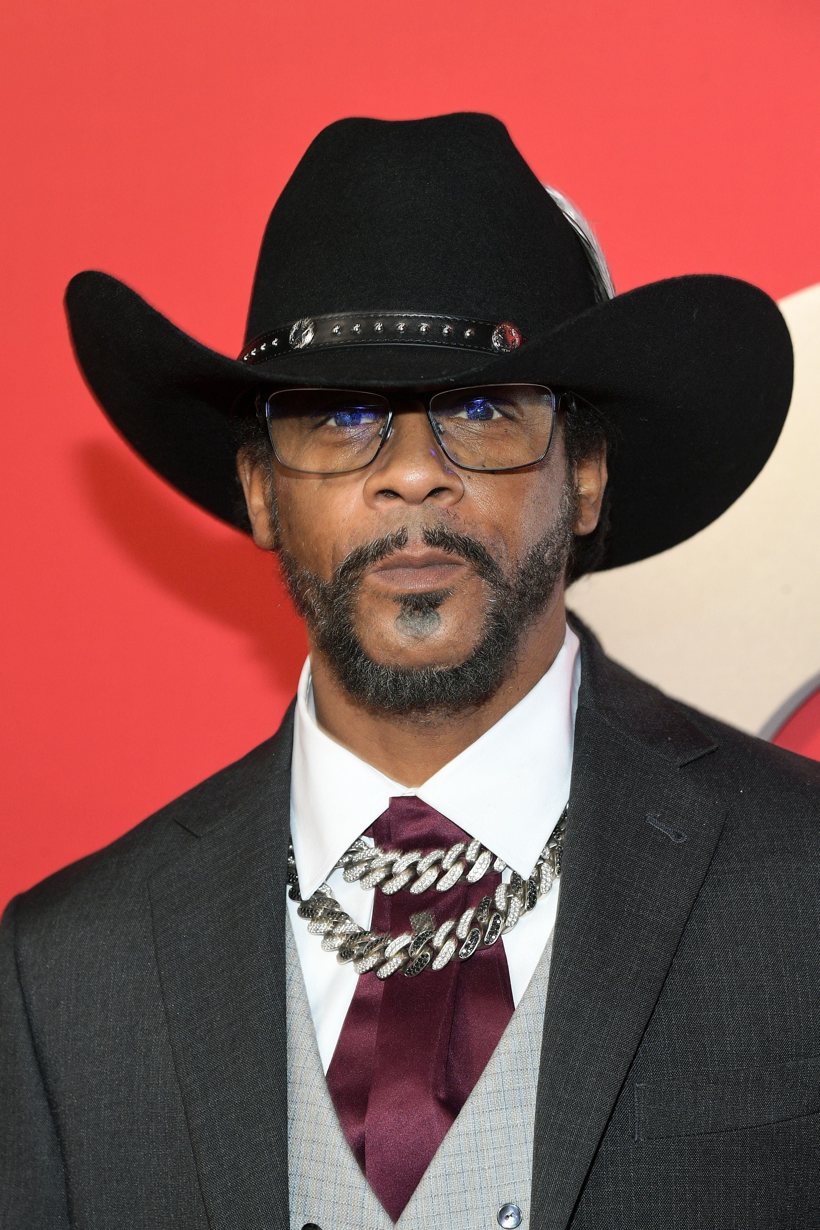Katt Williams in a cowboy hat wears glasses, a chain necklace, a formal suit, and a vest, posing on a red carpet