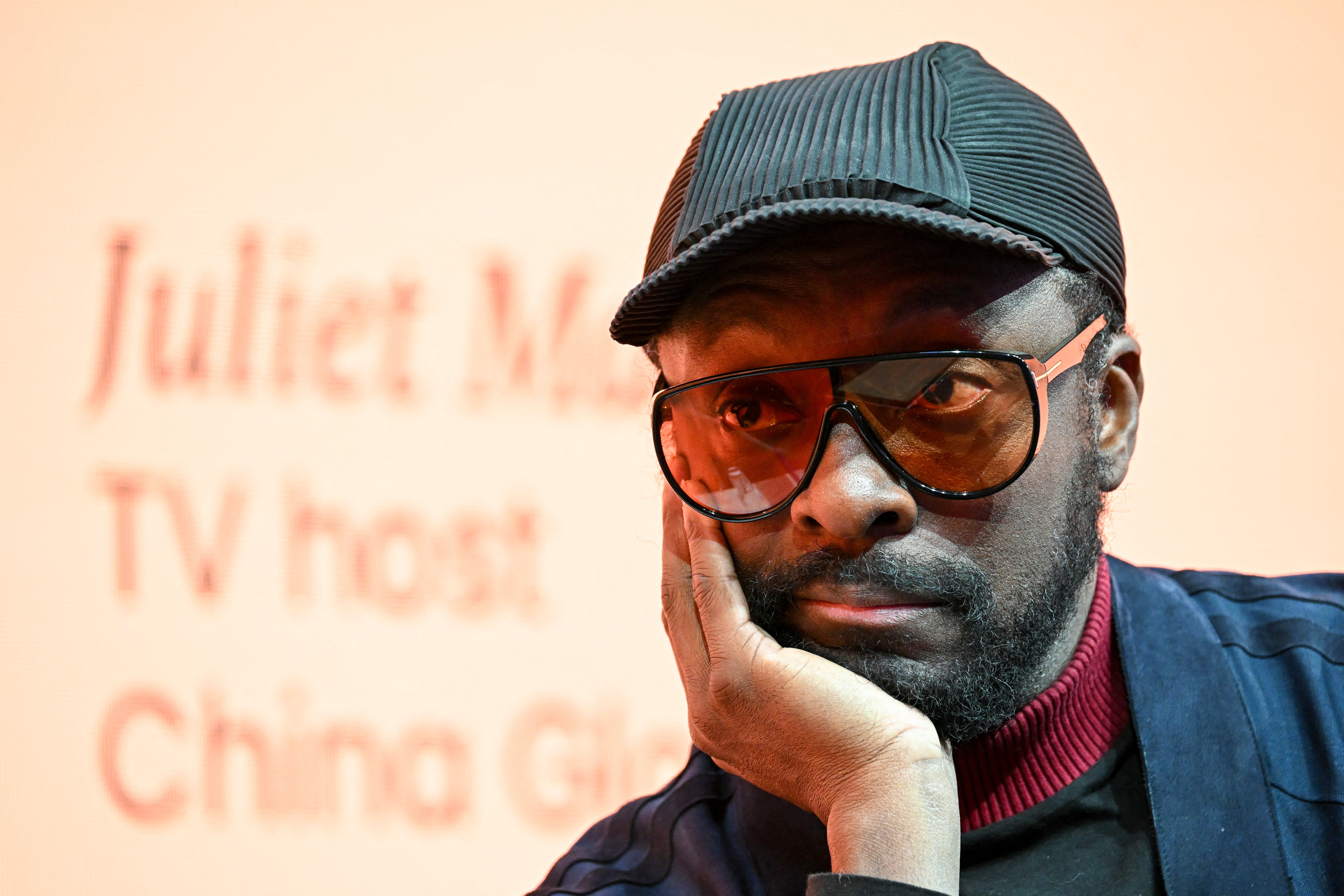 will.i.am wearing a pinstriped cap, tinted glasses, and a jacket with a red turtleneck, rests face on hand, looking thoughtful