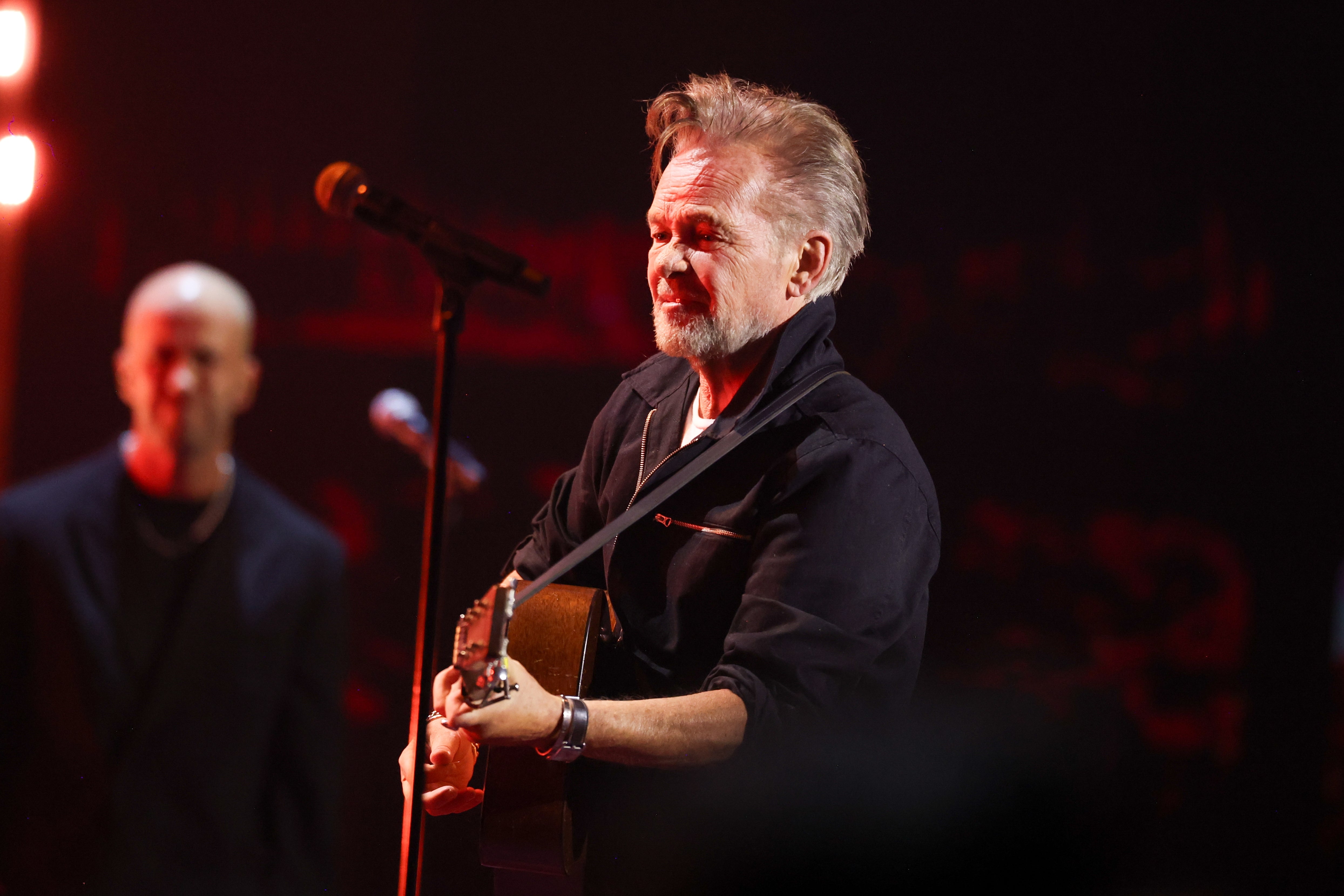 John Mellencamp performing on stage, playing guitar and singing into a microphone, wearing a casual jacket