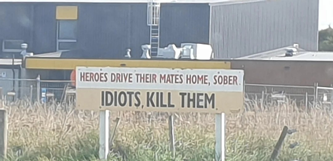 Sign reads: "HEROES DRIVE THEIR MATES HOME, SOBER. IDIOTS, KILL THEM," on roadside near industrial buildings