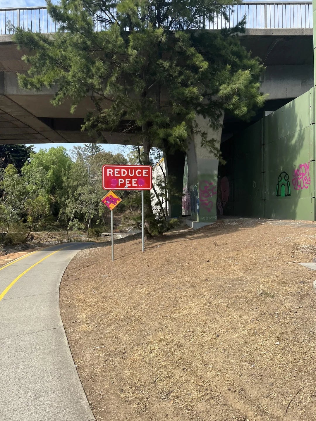 Sign near a pathway reads "REDUCE PEE" with altered text. Trees and an overpass are in the background