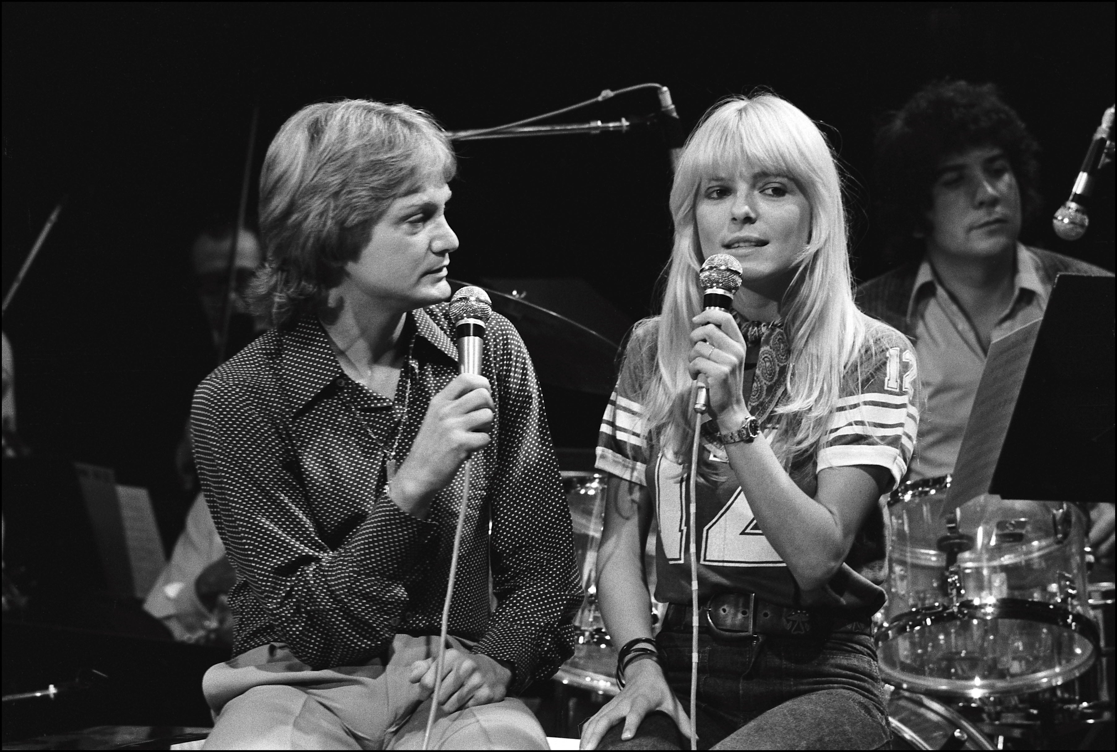 Two radical holding microphones, seated successful beforehand of a band. The idiosyncratic connected the near is wearing a patterned shirt; the idiosyncratic connected the close has agelong hairsbreadth and a sporty outfit