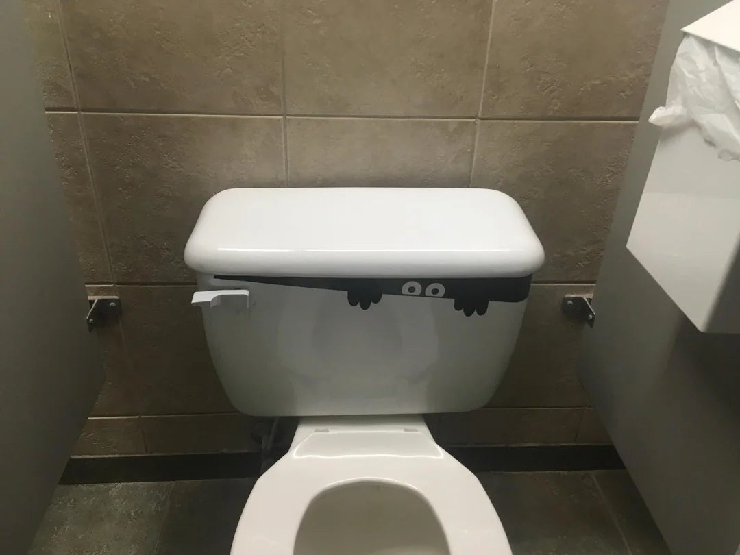 Toilet with a cartoon decal of eyes and hands peeking from behind the tank lid