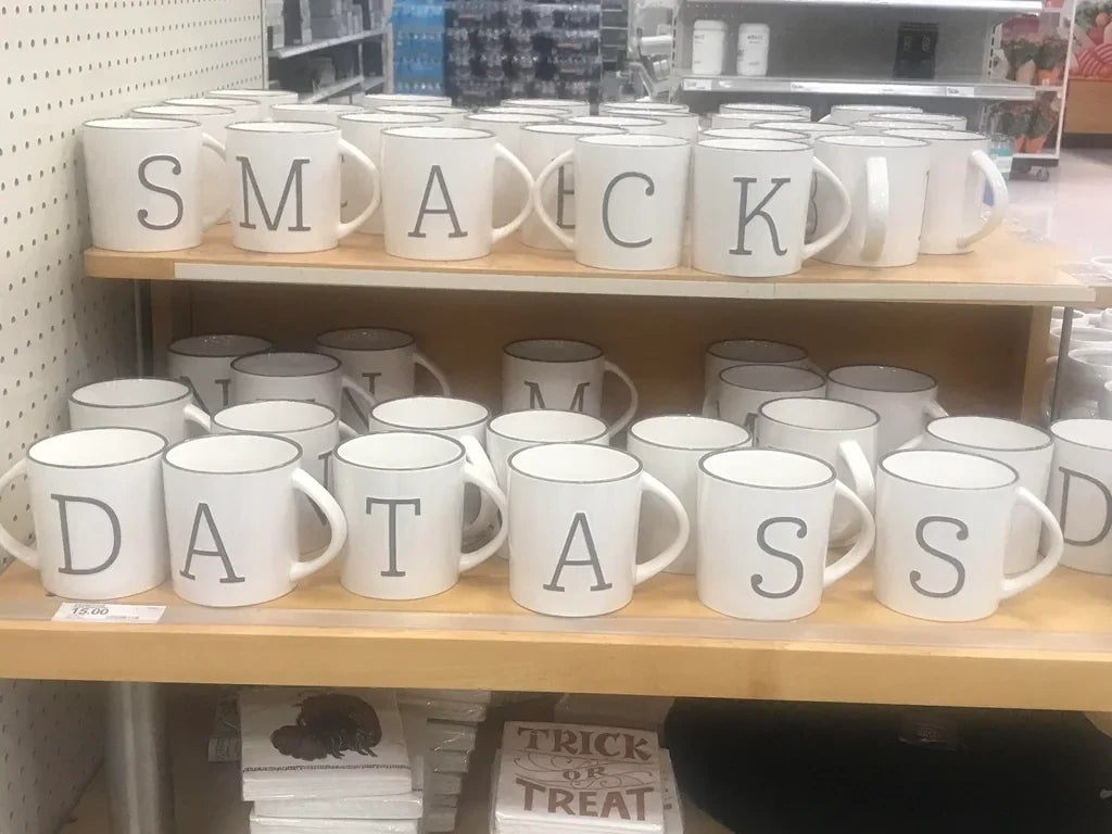 Mugs on a shelf display the letters "SMACK," "DAT," and "ASS," forming an unintended humorous phrase
