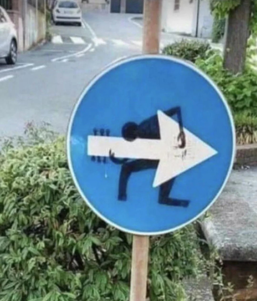 Traffic sign altered with art to show a person bending under the weight of a large arrow