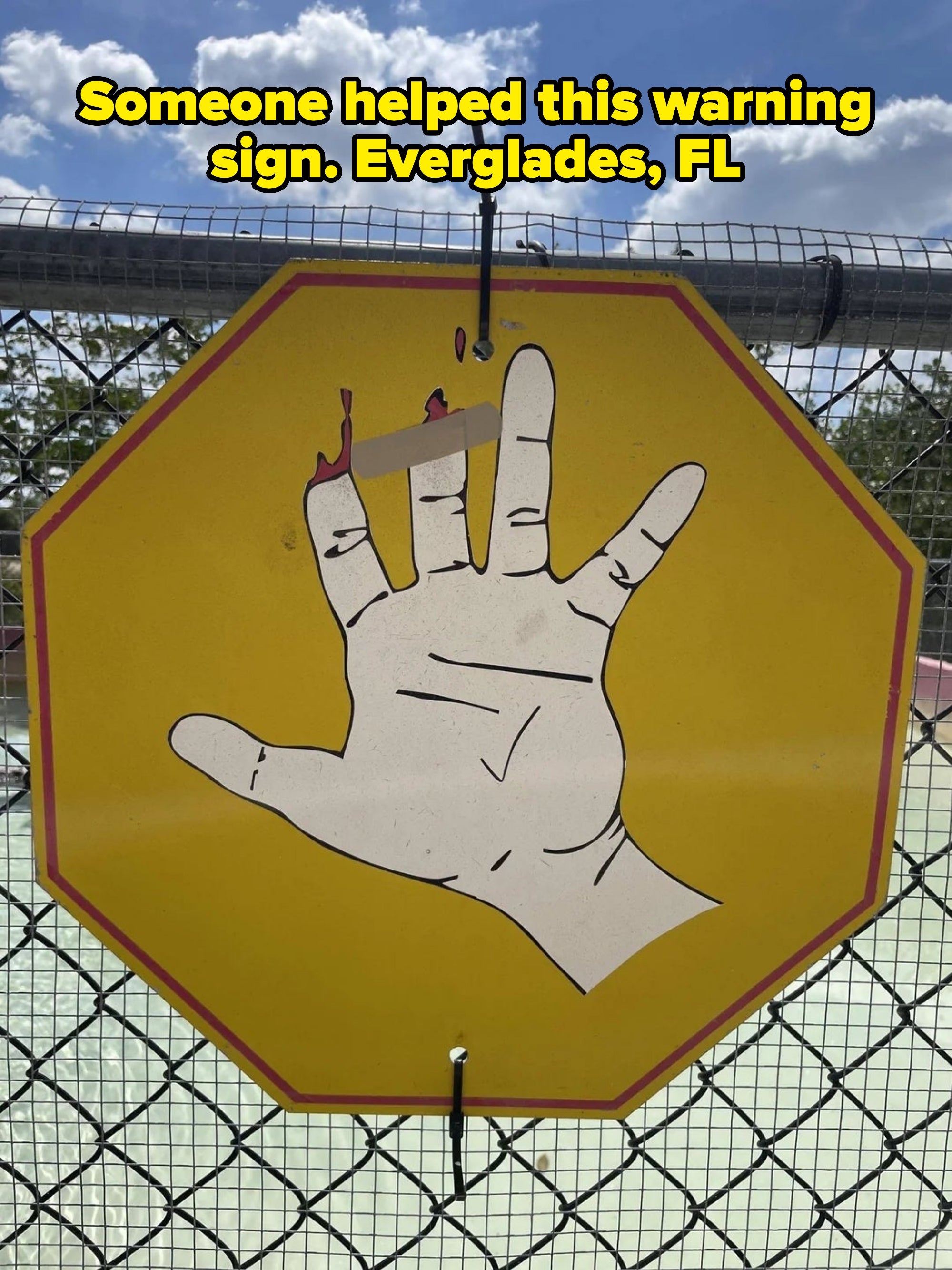 Warning sign with a hand symbol, missing two fingers, indicating a safety hazard behind a chain-link fence