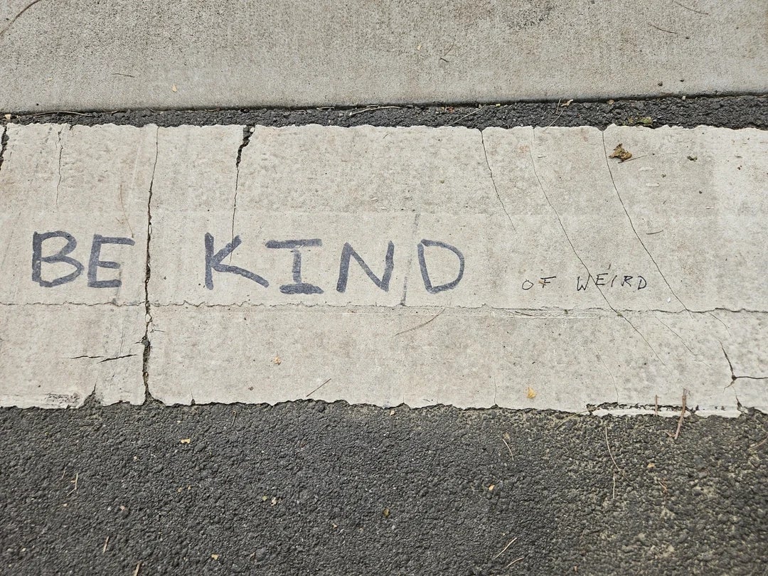 Pavement with the message "BE KIND of weird" written in large, uneven letters