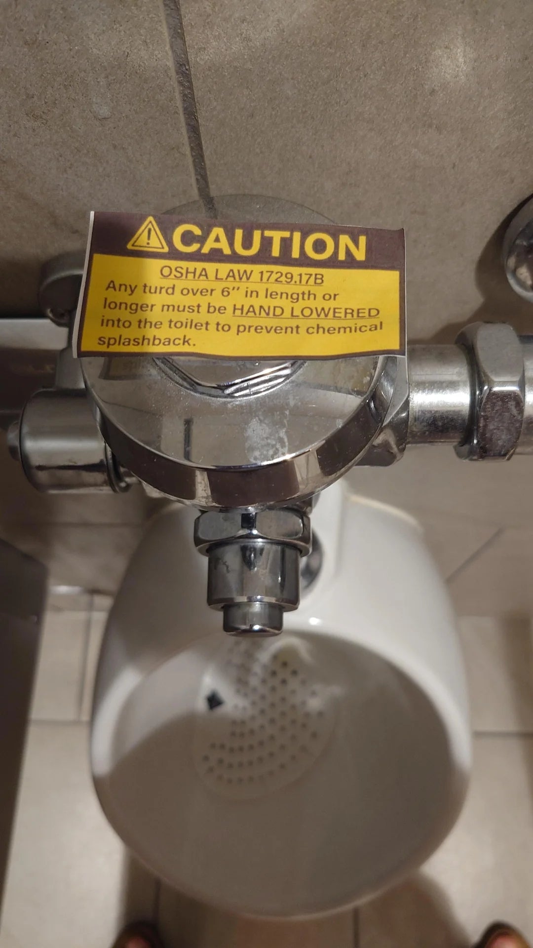 Warning sign above a urinal with text stating that OSHA law requires large waste to be hand-lowered into the toilet to prevent chemical splashback