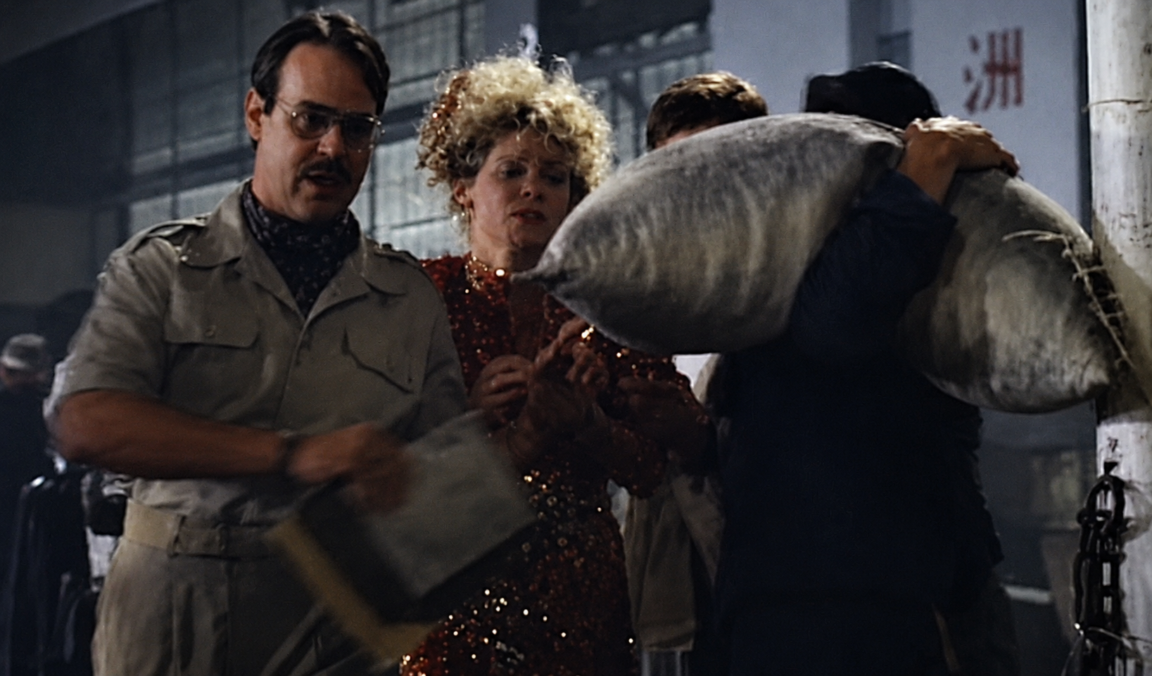 A group of people stand together, one holding a large sack. A person in a sparkly dress looks on. They appear to be on a movie set or film scene