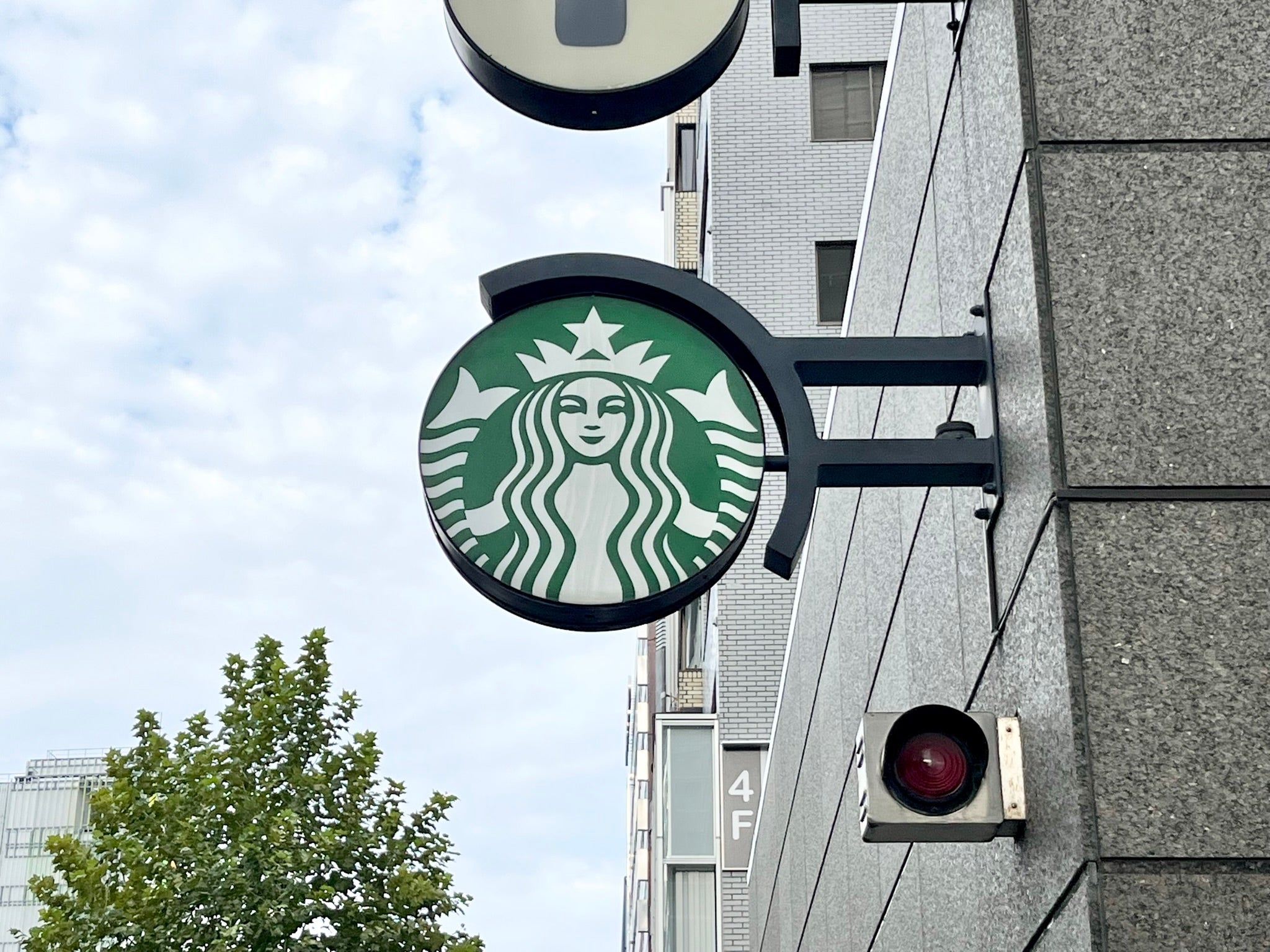 スターバックスの店舗看板が建物の外壁に取り付けられている様子。背景には空と木が見える。