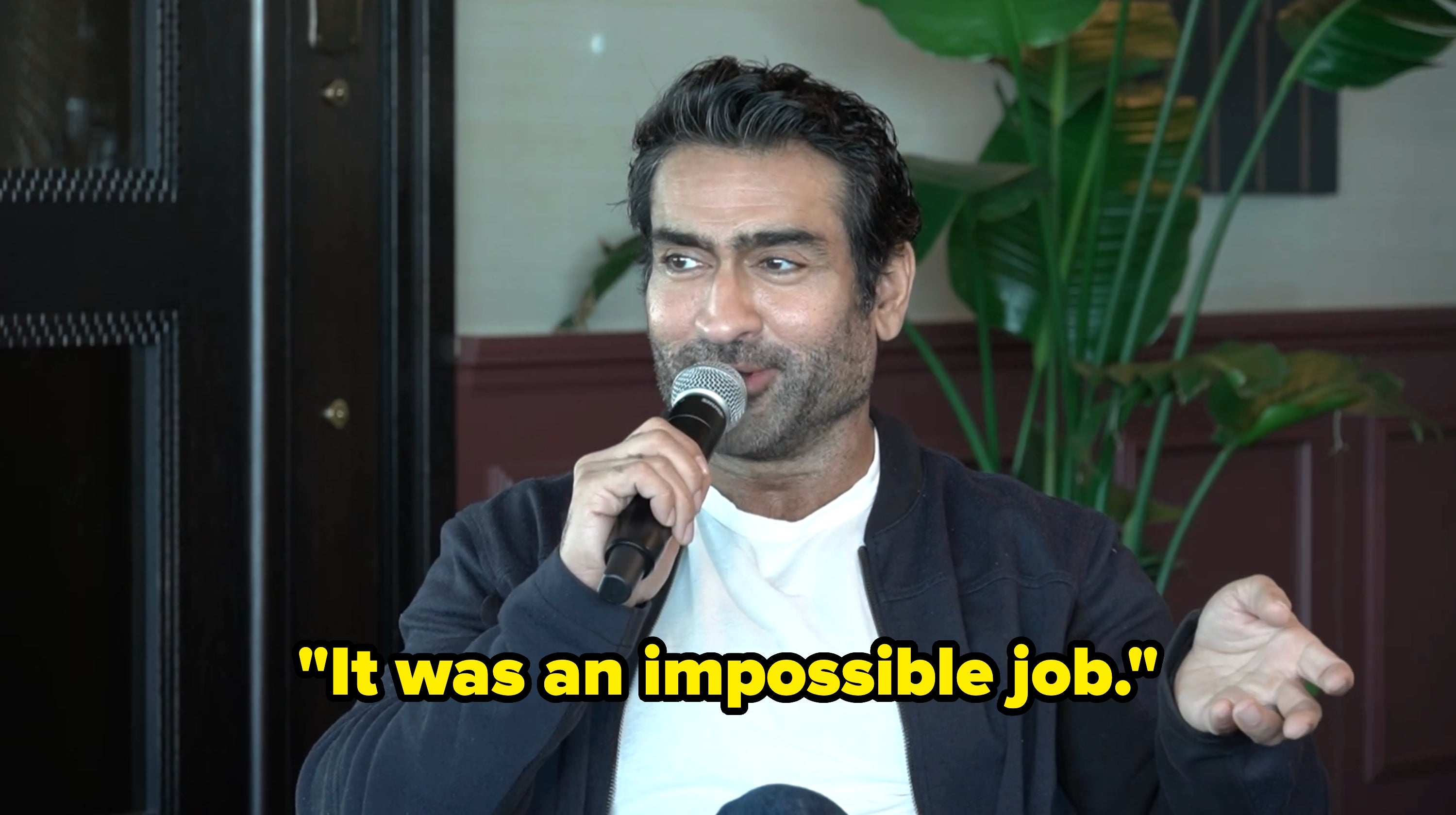 Person sitting and speaking into a microphone, gesturing with 1  hand, with a works  successful  the background