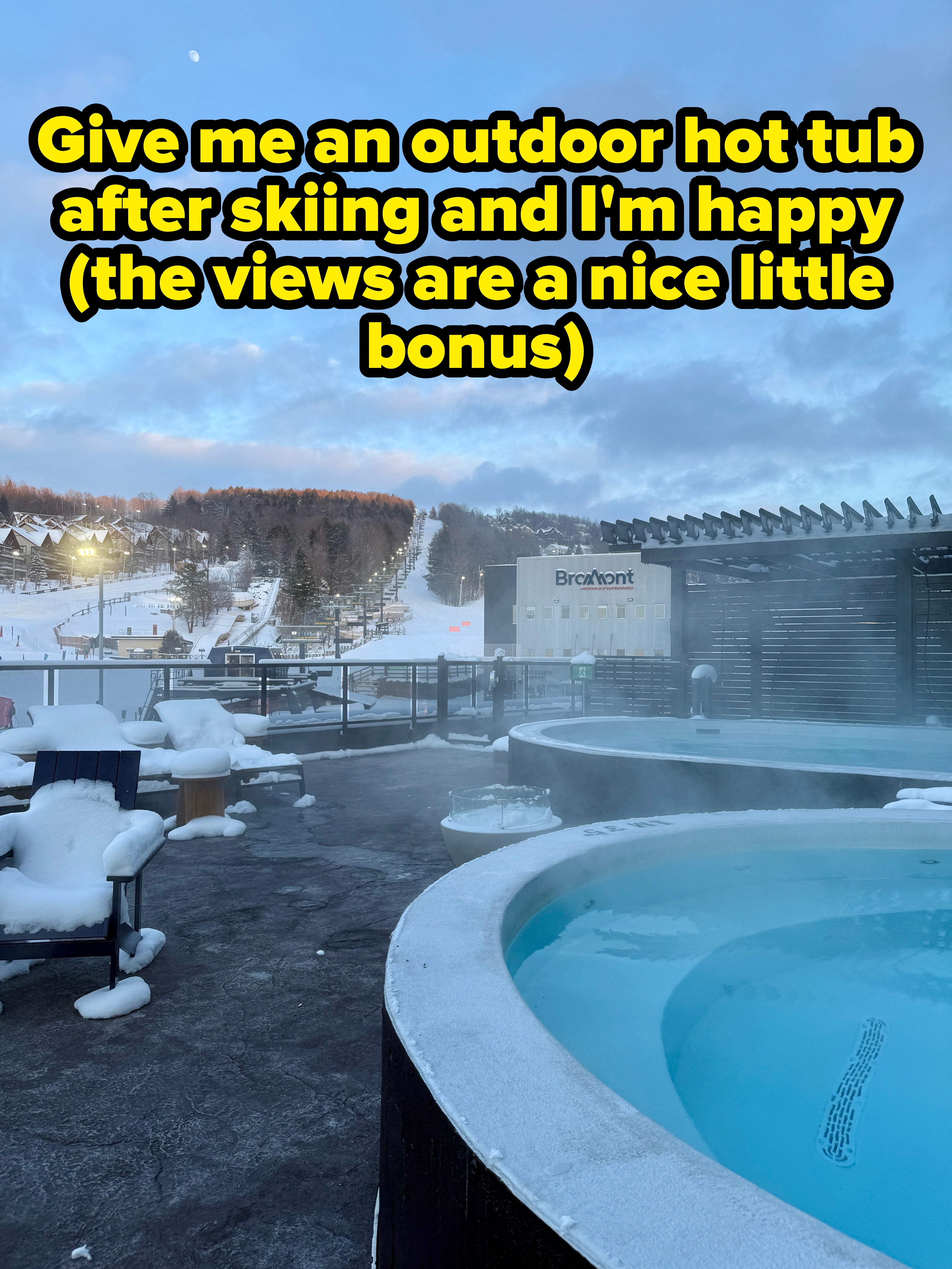 Hot tub with snow, overlooking a skis edifice during dusk, steam rising. Relaxing wintertime getaway atmosphere
