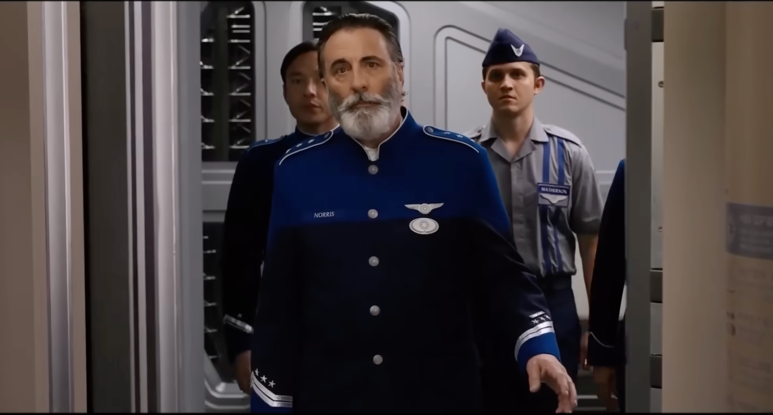 A man in a blue military uniform with a beard walks in a futuristic setting, followed by others in similar attire