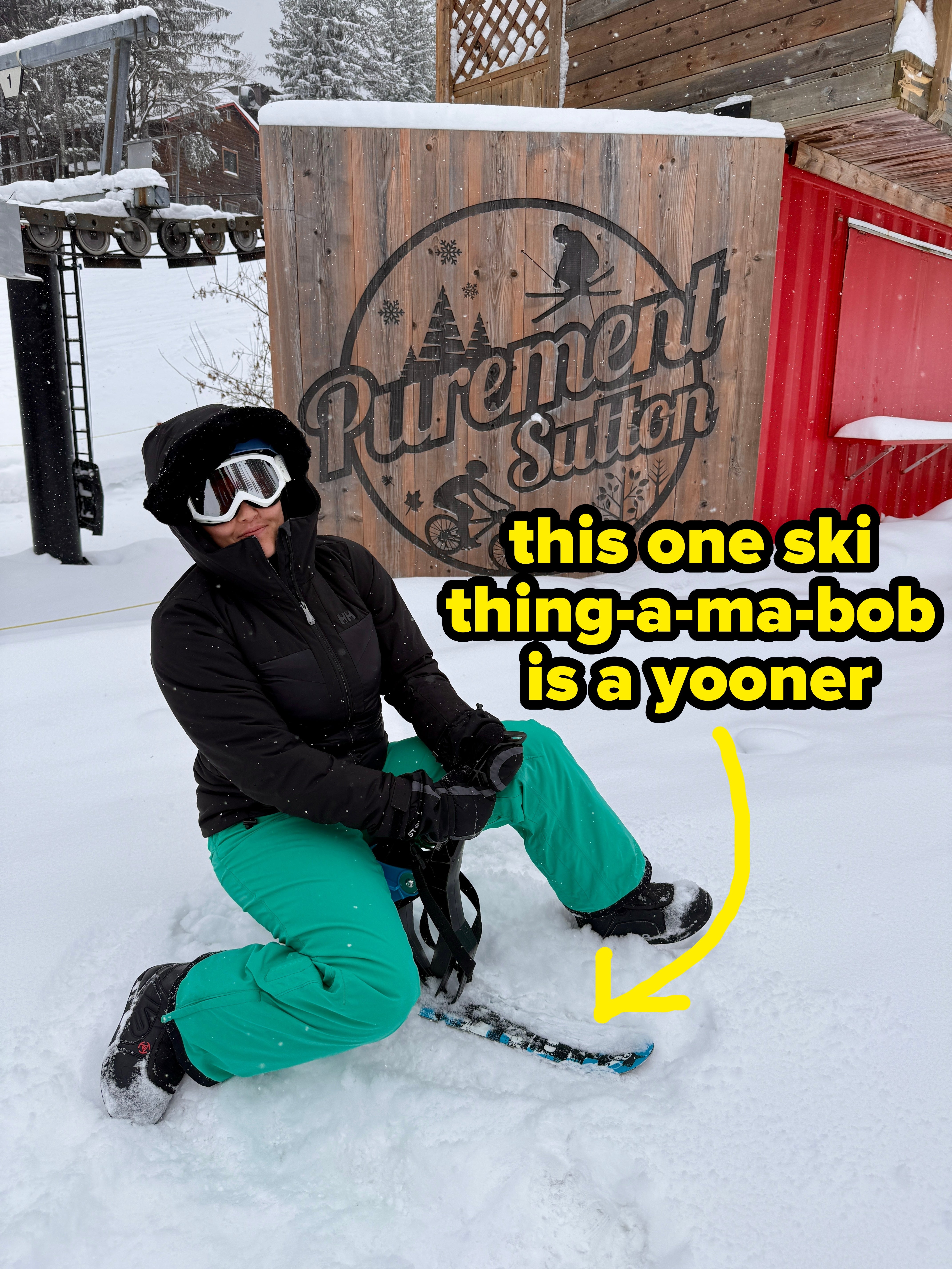 Person successful skis cogwheel kneels successful snowfall adjacent a woody motion speechmaking "Purement Sutton" with snowy trees and buildings successful the background