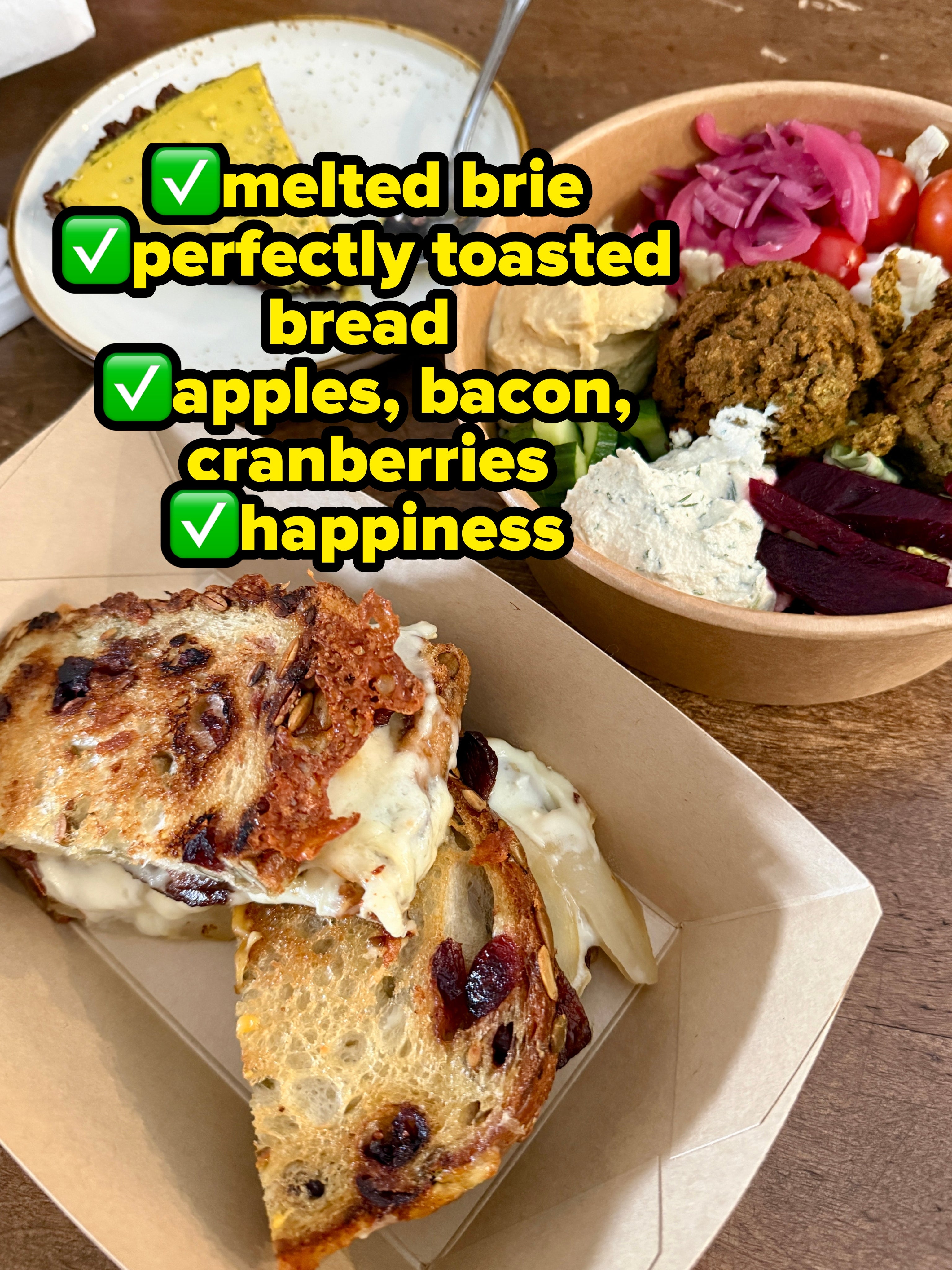 A grilled food sandwich, a portion of quiche, and a vessel with falafel, hummus, tomatoes, and vegetables are connected a woody table