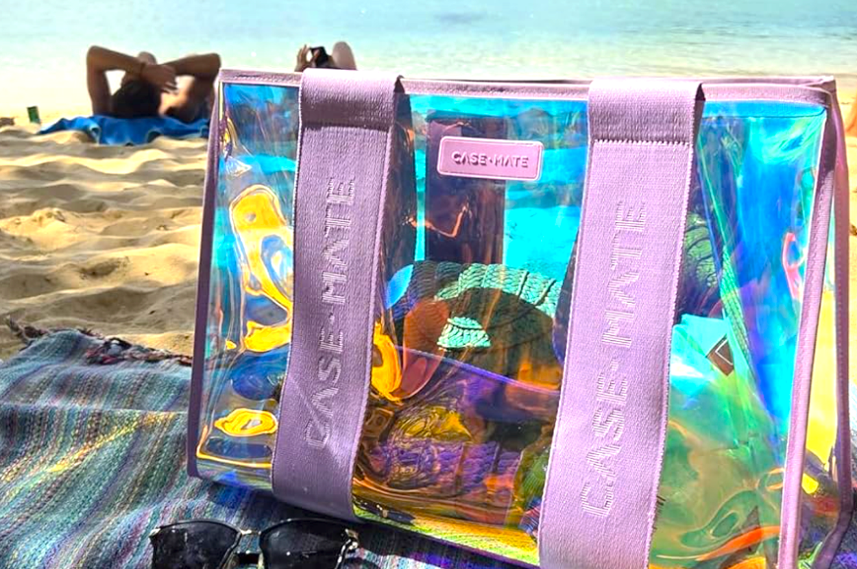 Clear beach bag with pink straps on a sandy beach, near sunglasses, sunblock, and a straw hat, with people lounging by the sea