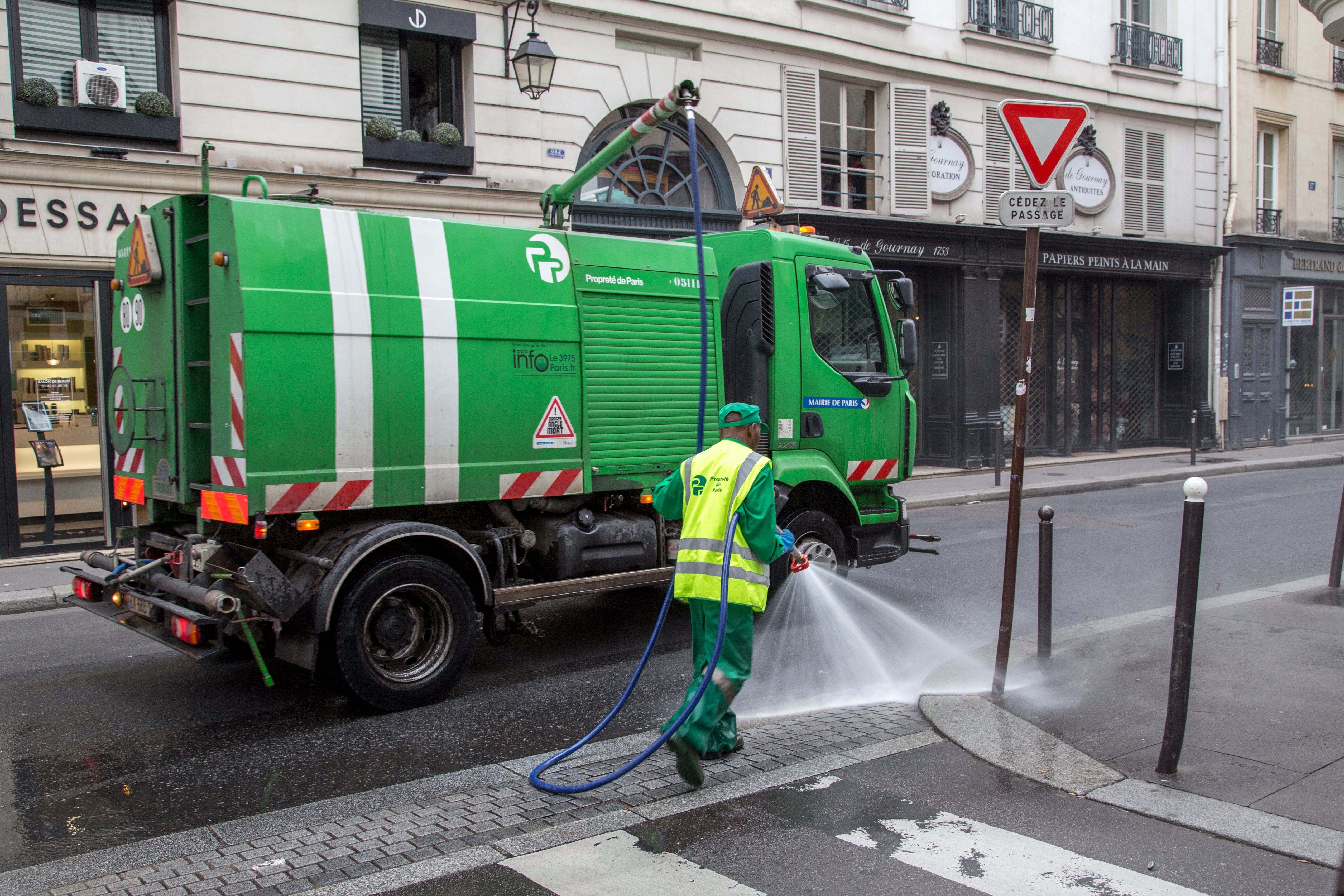 Street cleaner successful protective cogwheel uses a hose to lavation a Parisian street. A greenish cleaning motortruck is parked nearby. Shops are disposable successful the background