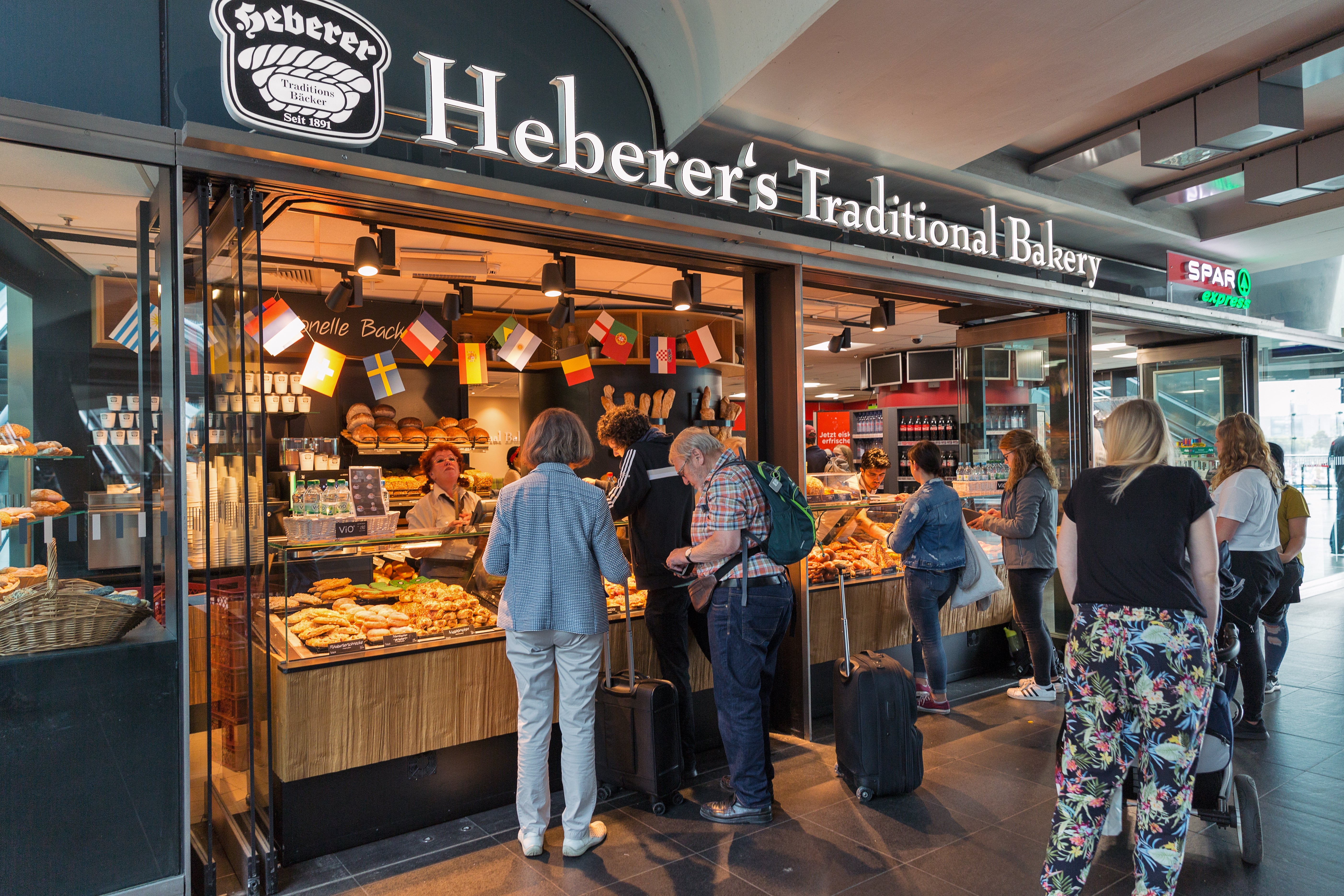 People queue astatine Heberer's Traditional Bakery successful an airport, waiting to acquisition pastries and bread. Flags are displayed supra the counter