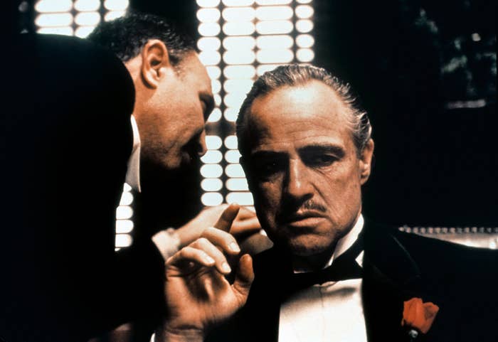 A antheral   successful  a tuxedo with a roseate  lapel listens intently to different  antheral   whispering successful  his ear, acceptable   against a dimly lit, ceremonial  background