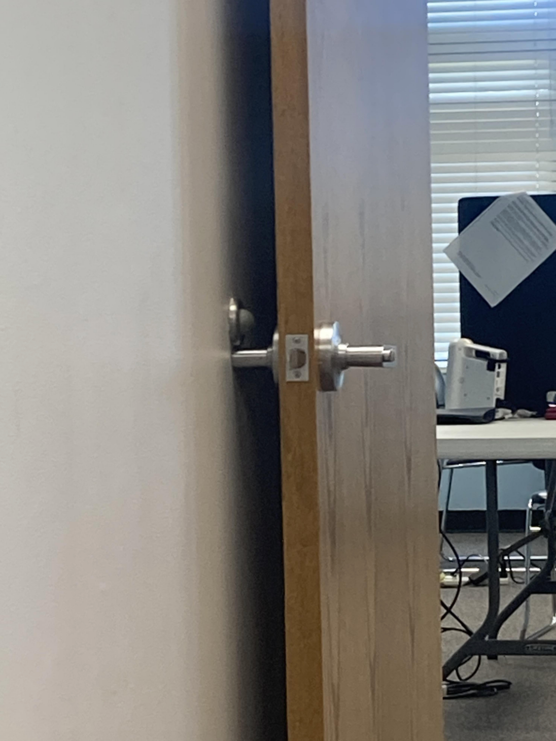 Sebuah pintu terbuka sebagian, memperlihatkan ruang kantor dengan meja dan peralatan komputer. Sinar matahari menyaring melalui tirai di latar belakang