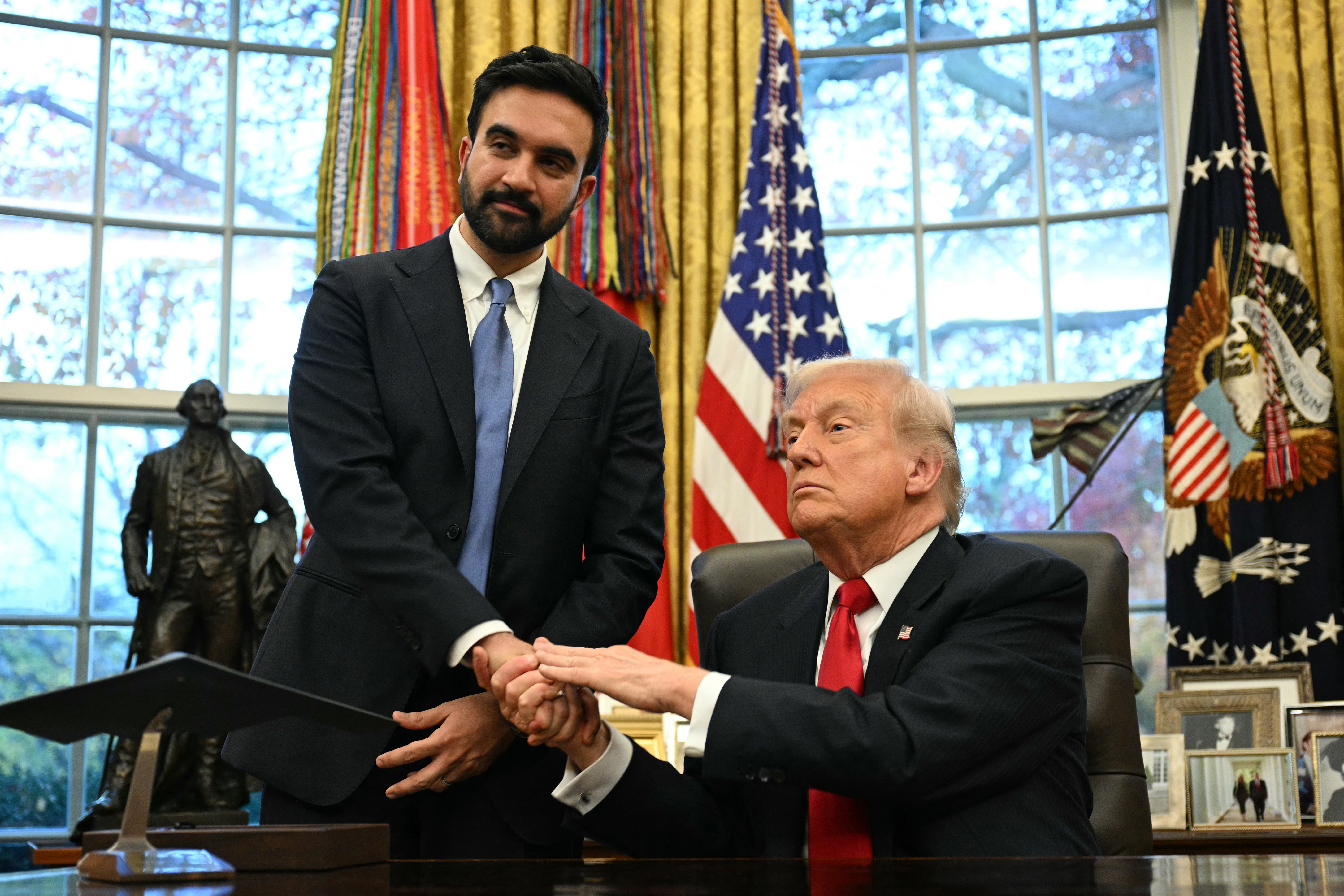 Two radical   successful  a ceremonial  mounting  shingle  hands; 1  sits astatine  a desk, the different   stands beside, some  successful  suits. American flags are disposable   successful  the background