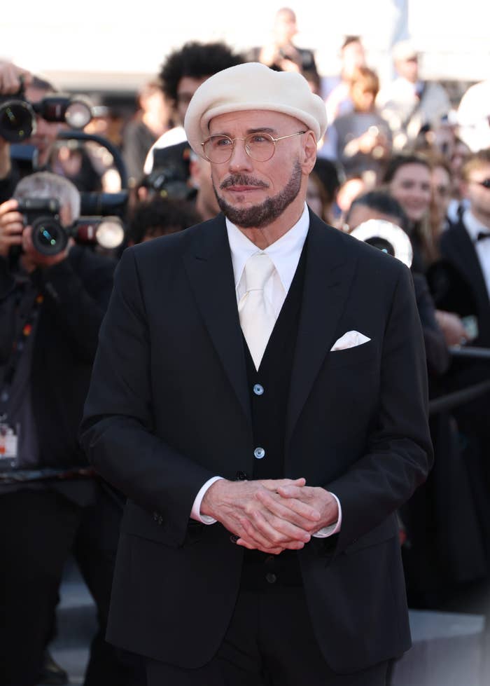 Person in a suit with a beret and glasses stands on a red carpet, surrounded by photographers