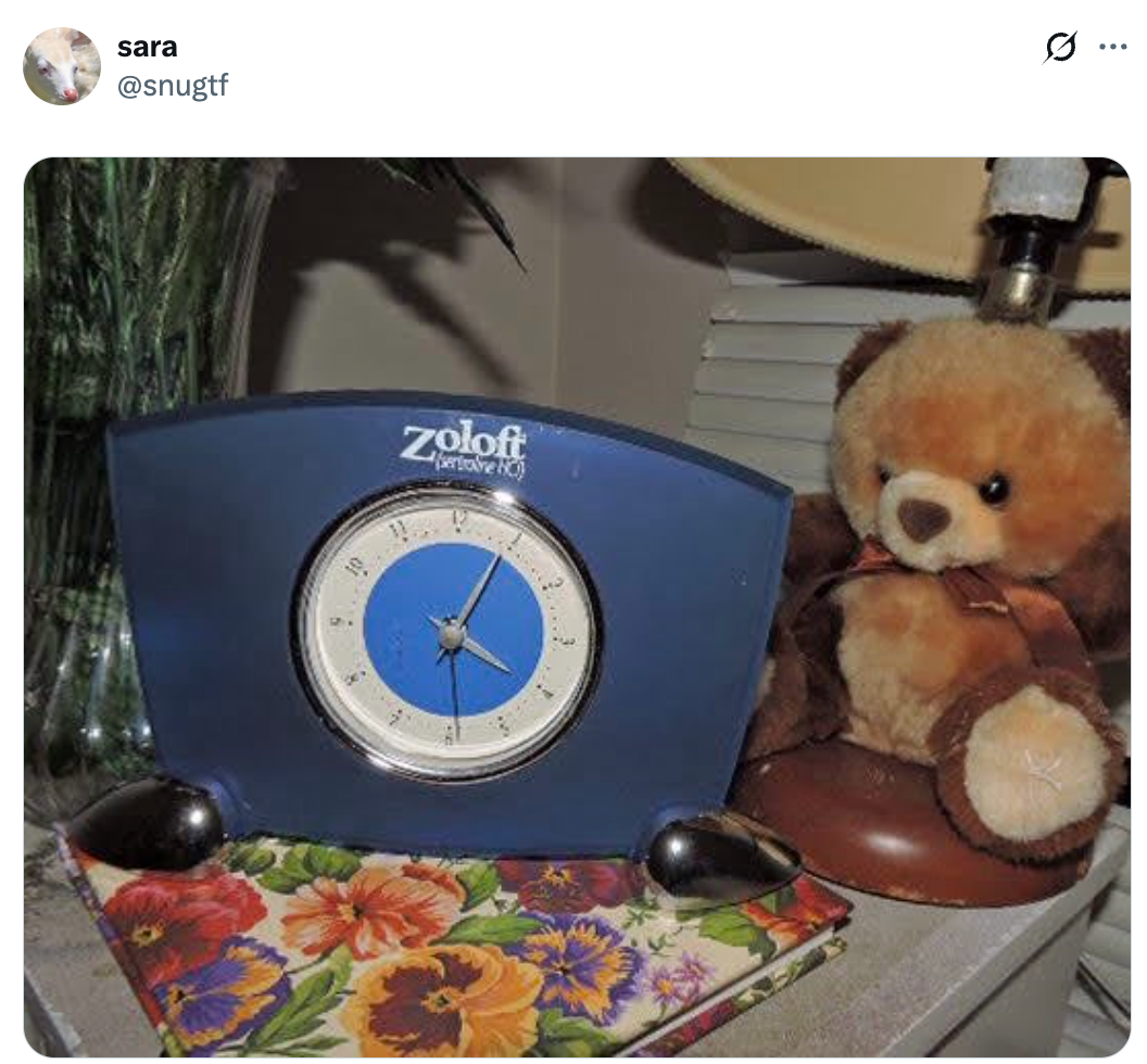 En klokke med Zoloft-merke, en teddybjørn og en blomstermønstret notatbok ligger på et bord under en lampe