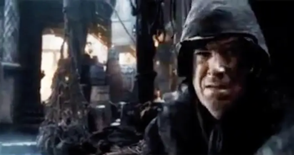 Scene from a dark fantasy film showing a hooded character with a tense expression, surrounded by mysterious and shadowy props