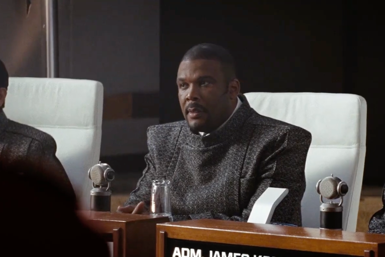 A person in a futuristic setting speaks at a conference table, wearing a sleek modern suit, suggesting a sci-fi or action film scene