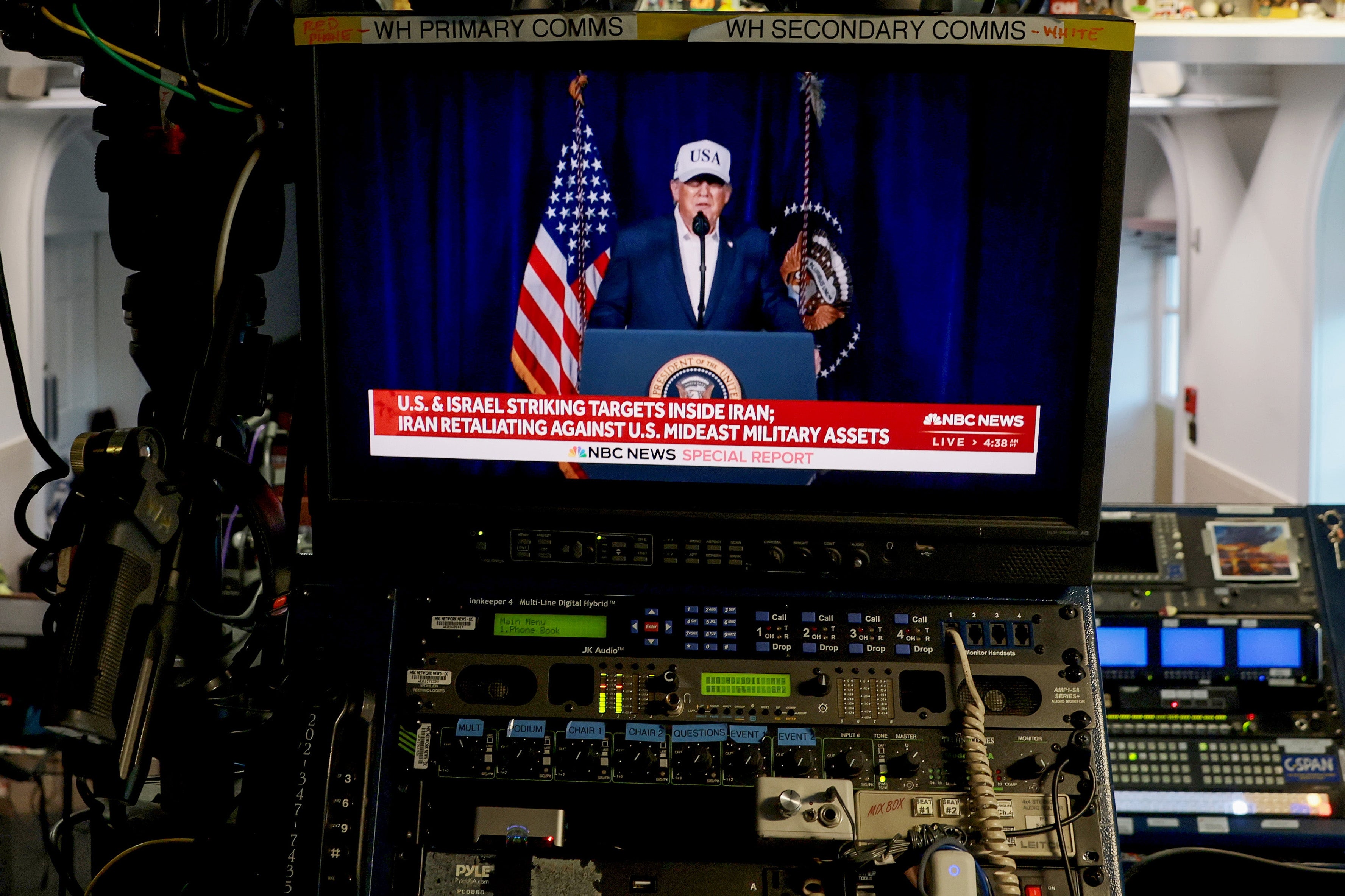 En person som har på seg en dress og et "USA" cap snakker på et podium med det amerikanske flagget og presidentens segl, vist på en kringkastingsmonitor i et kontrollrom
