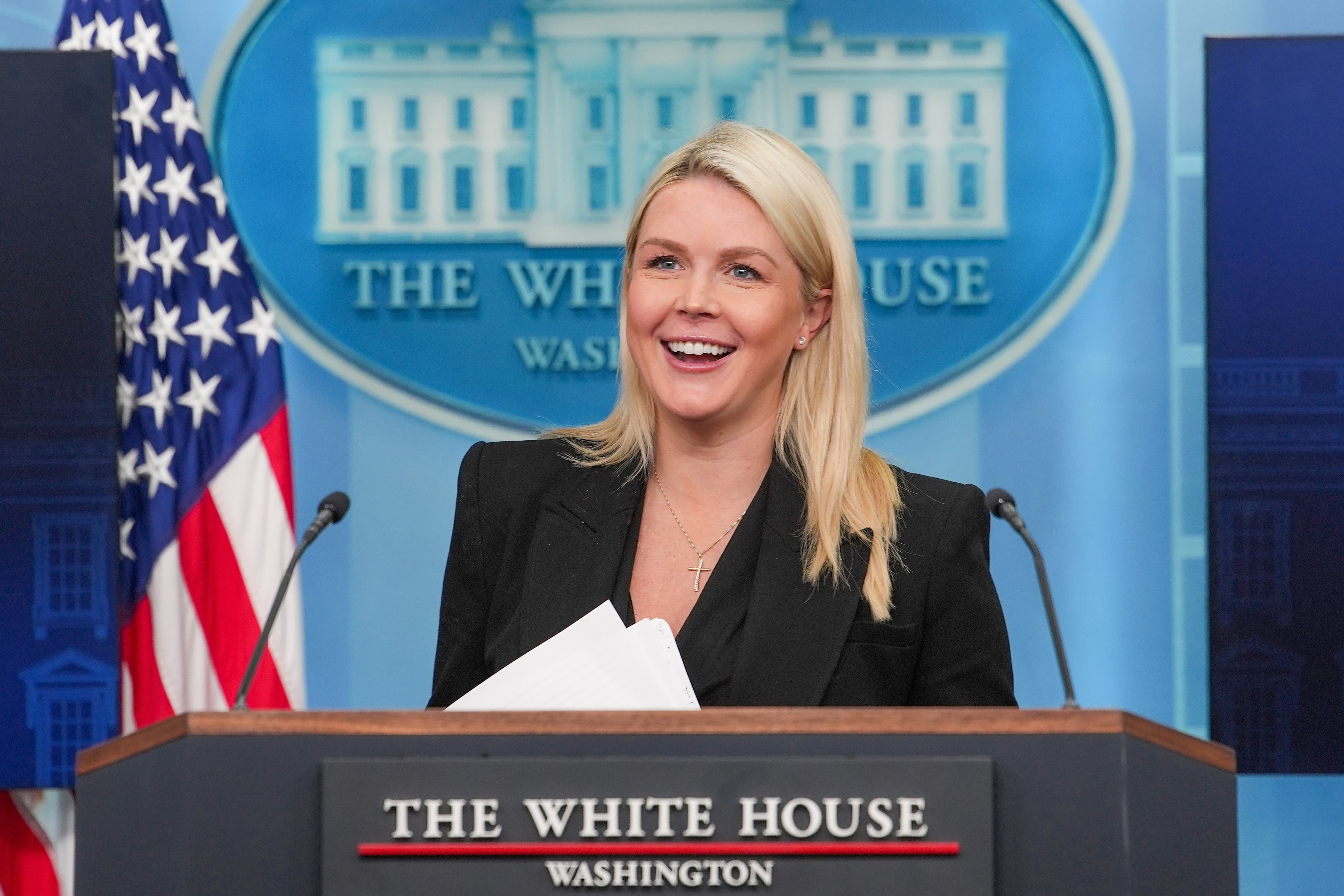 Person som snakker på podiet i Det hvite hus, smiler, holder papirer. Amerikansk flagg og Det hvite hus-emblem i bakgrunnen