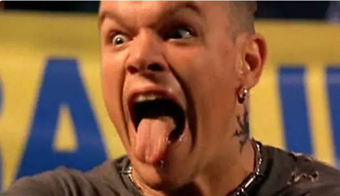 A person with a surprised expression sticks their tongue out, wearing earrings and a neck tattoo, set against a blurred background