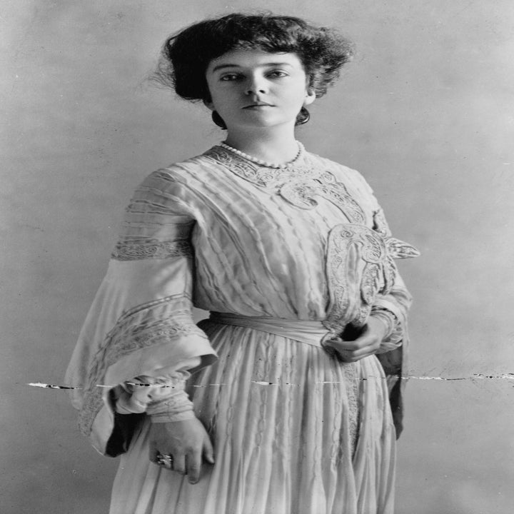 A woman from the early 1900s wearing a long, elegant dress with lace detailing, posing formally in a studio setting