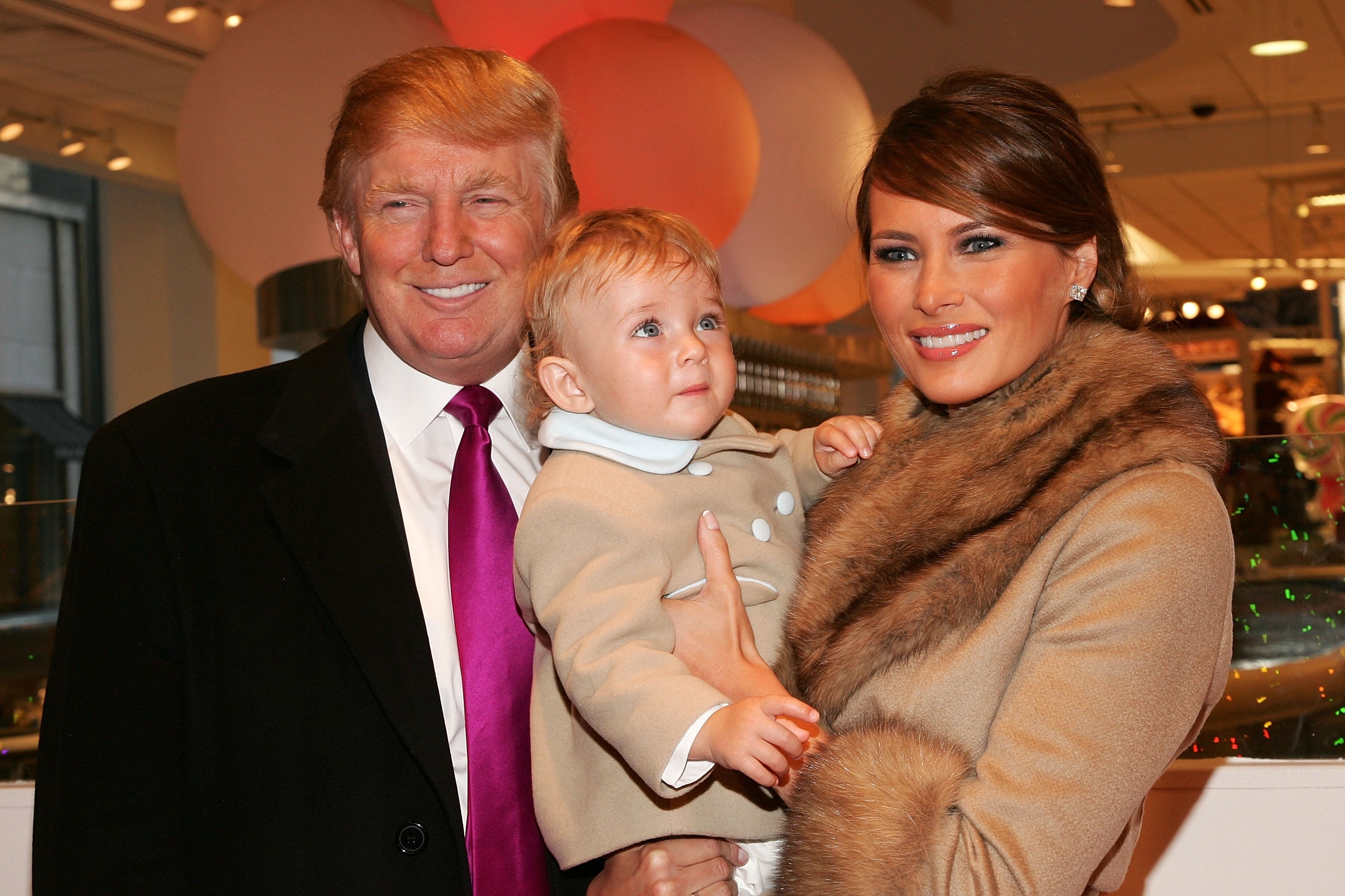 A antheral successful a suit and necktie and a pistillate successful a fur-trimmed overgarment grin portion holding a toddler successful ceremonial deterioration astatine an indoor event
