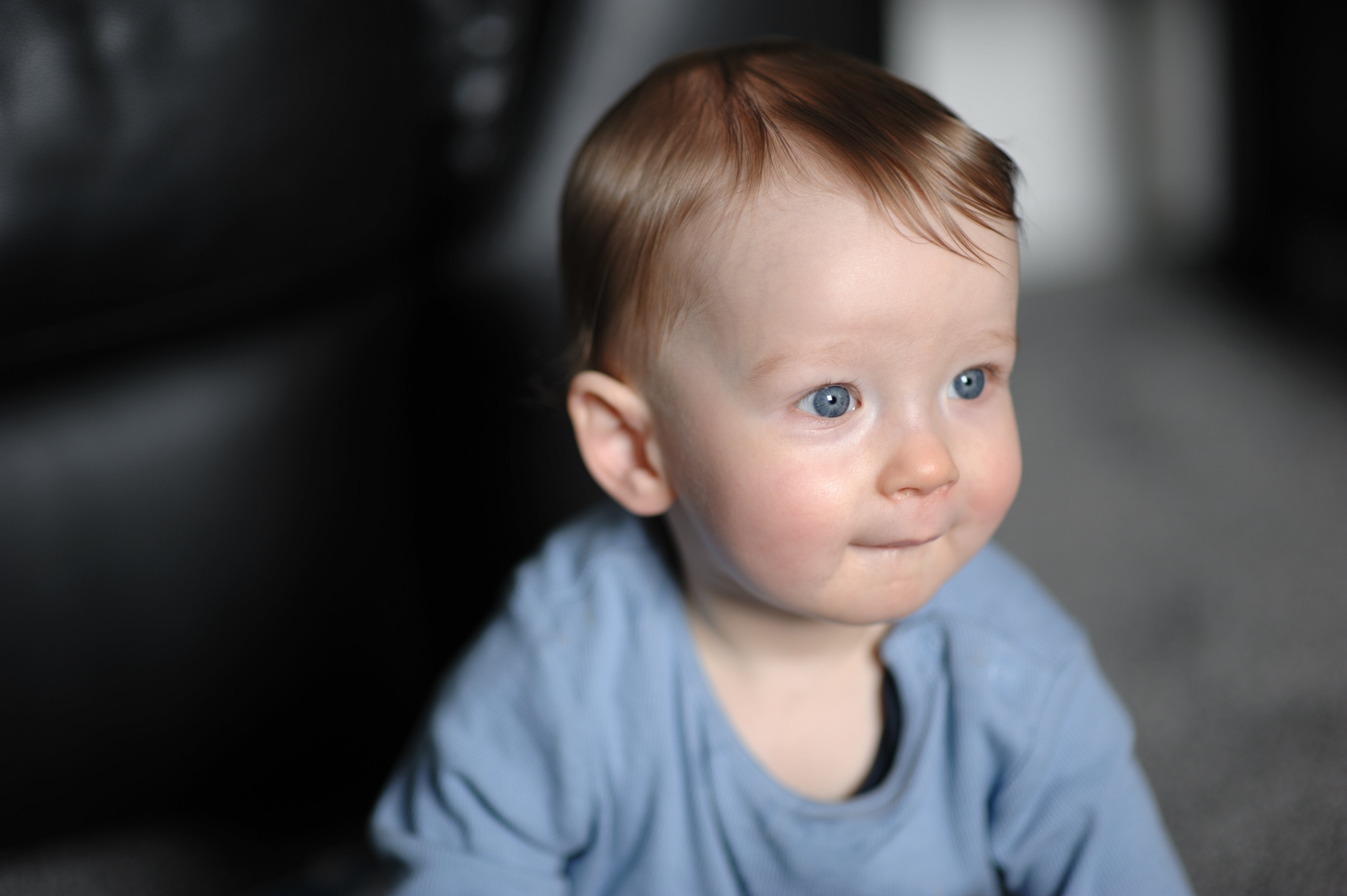 Baby with airy hairsbreadth and bluish eyes, wearing a brushed onesie, sits attentively, showing a hint of a smile