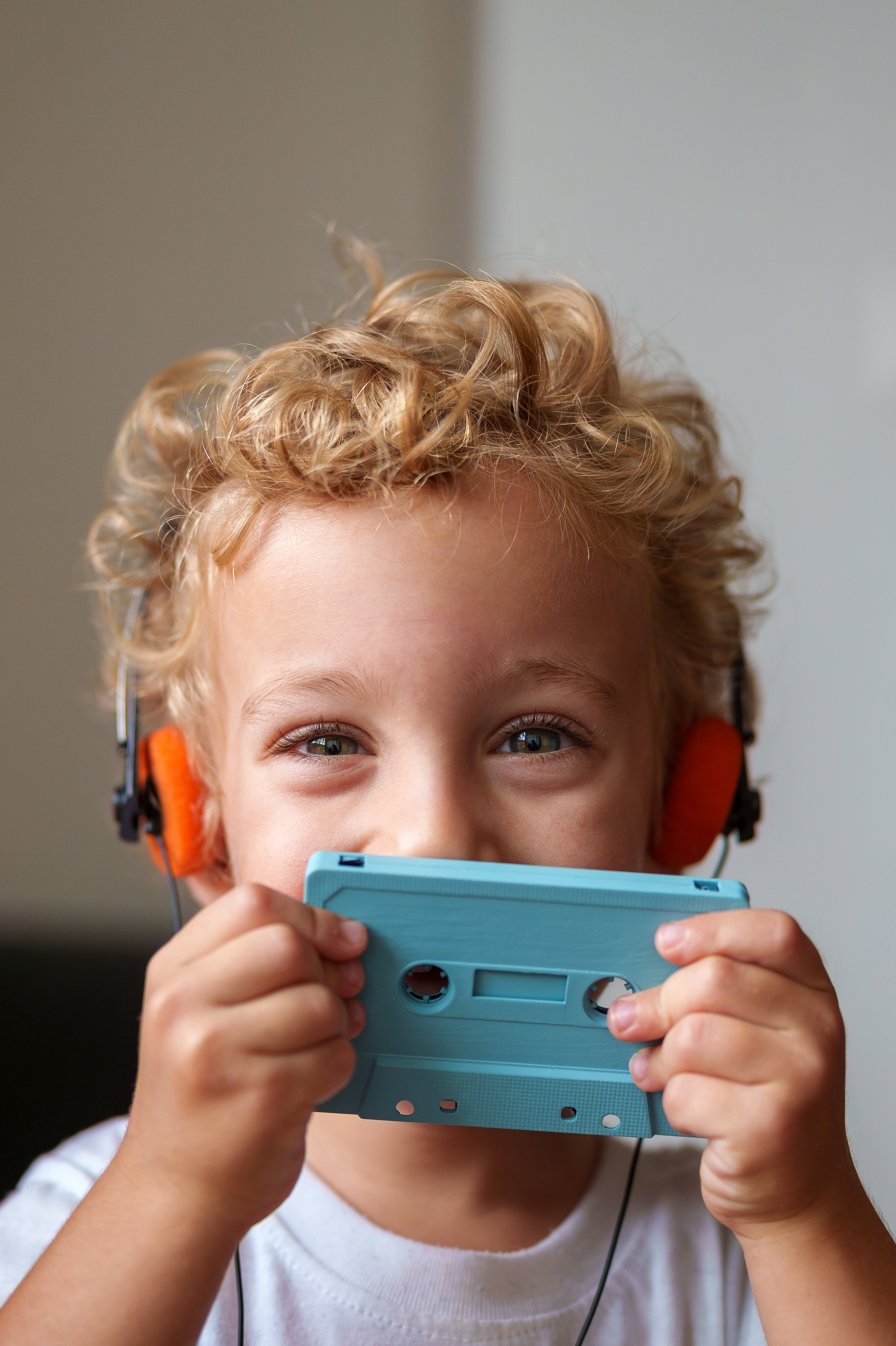 Child with curly hairsbreadth holds a bluish cassette portion up to their look portion wearing headphones, smiling playfully