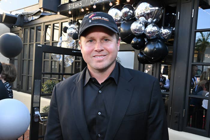 Person in a black suit and cap at an outdoor event with decorative balloons in the background