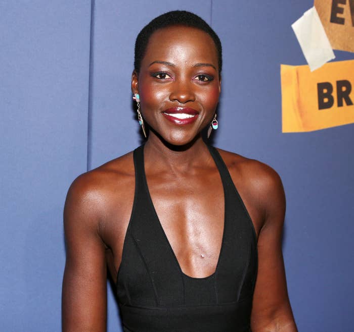 Person in a sleeveless black dress smiles at an event, standing against a backdrop with text partially visible