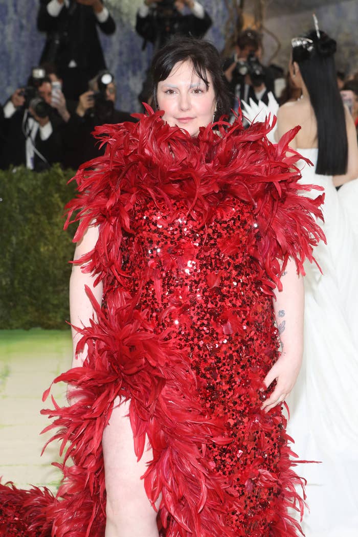 Person wearing a gown covered in large feathers and sequins on a red carpet, with photographers in the background