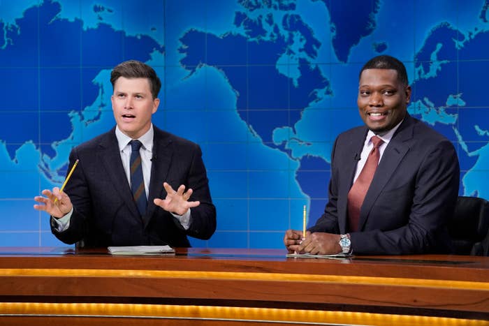 Two TV hosts in suits sit behind a news desk, holding pencils, in front of a world map backdrop