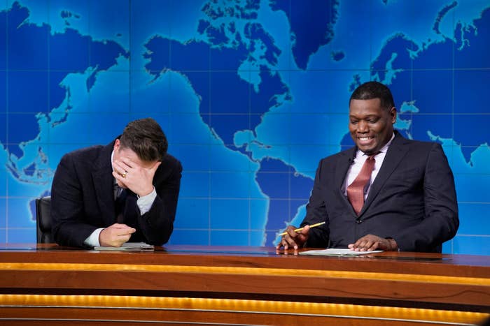 Two men sit at a news desk, one laughing with his head in his hand, the other smiling and holding a pen