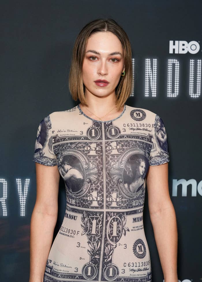 Person on a red carpet wearing a dress featuring a dollar bill print, standing in front of a media backdrop