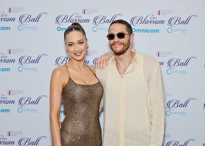Two people smiling on a Blossom Ball red carpet; one in a shimmering gown, the other in a casual shirt with sunglasses